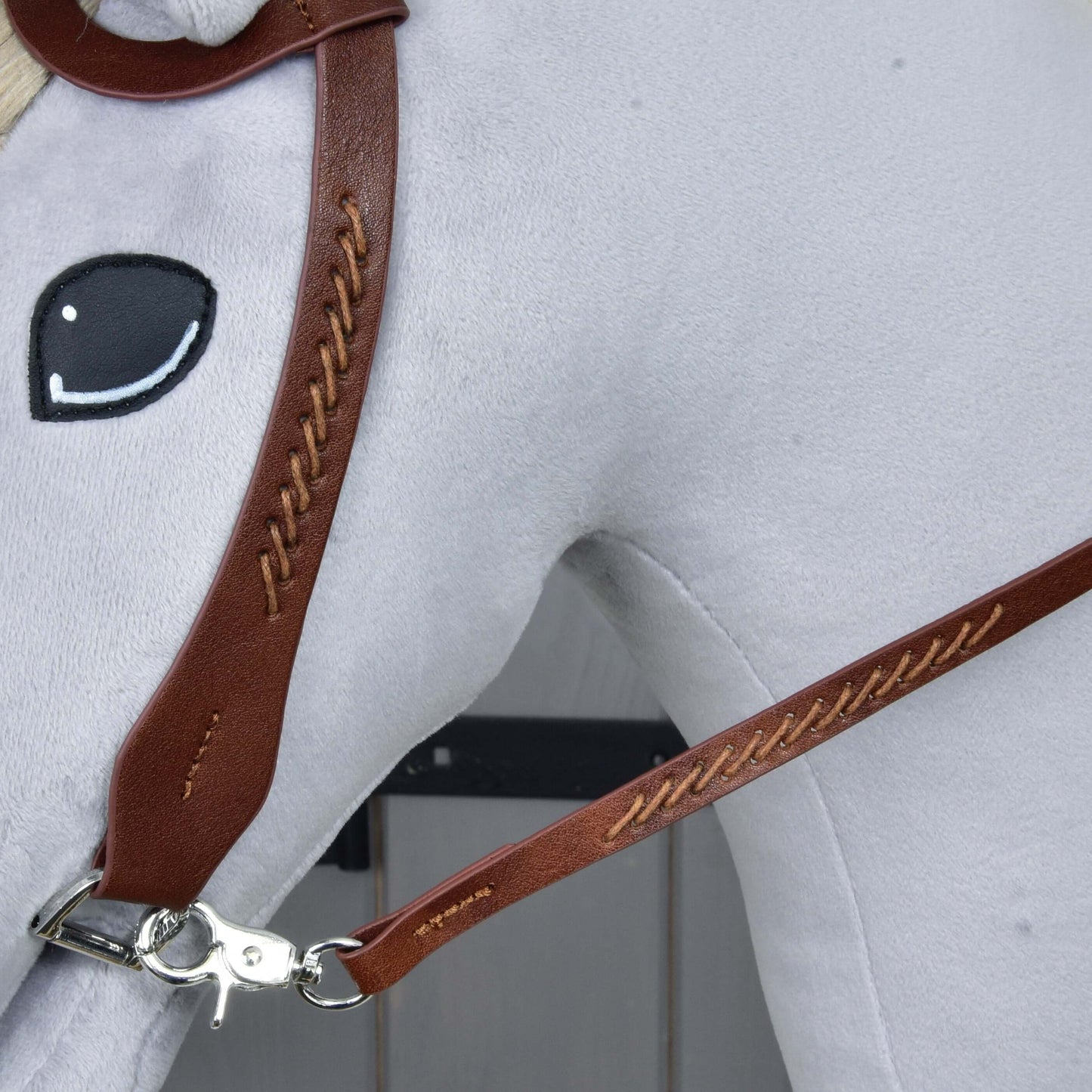 Close-up of a Hobby Horse LarDen plush horse head wearing the Complete Western bridle One-Ear with brown leather, detailed stitching, split reins, metal clip, and a simple black and white embroidered eye on a light gray background.