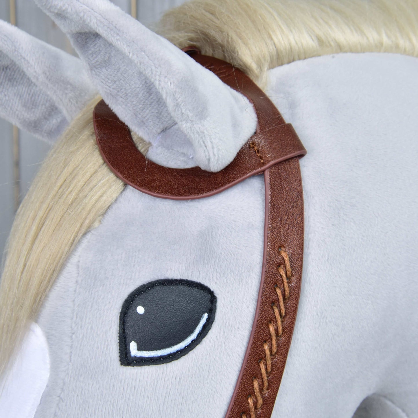 A close-up of a Hobby Horse LarDen plush with light gray fur, cream mane, black and white embroidered eye, wearing the Complete Western bridle One-Ear with split reins and a sliding ear loop.