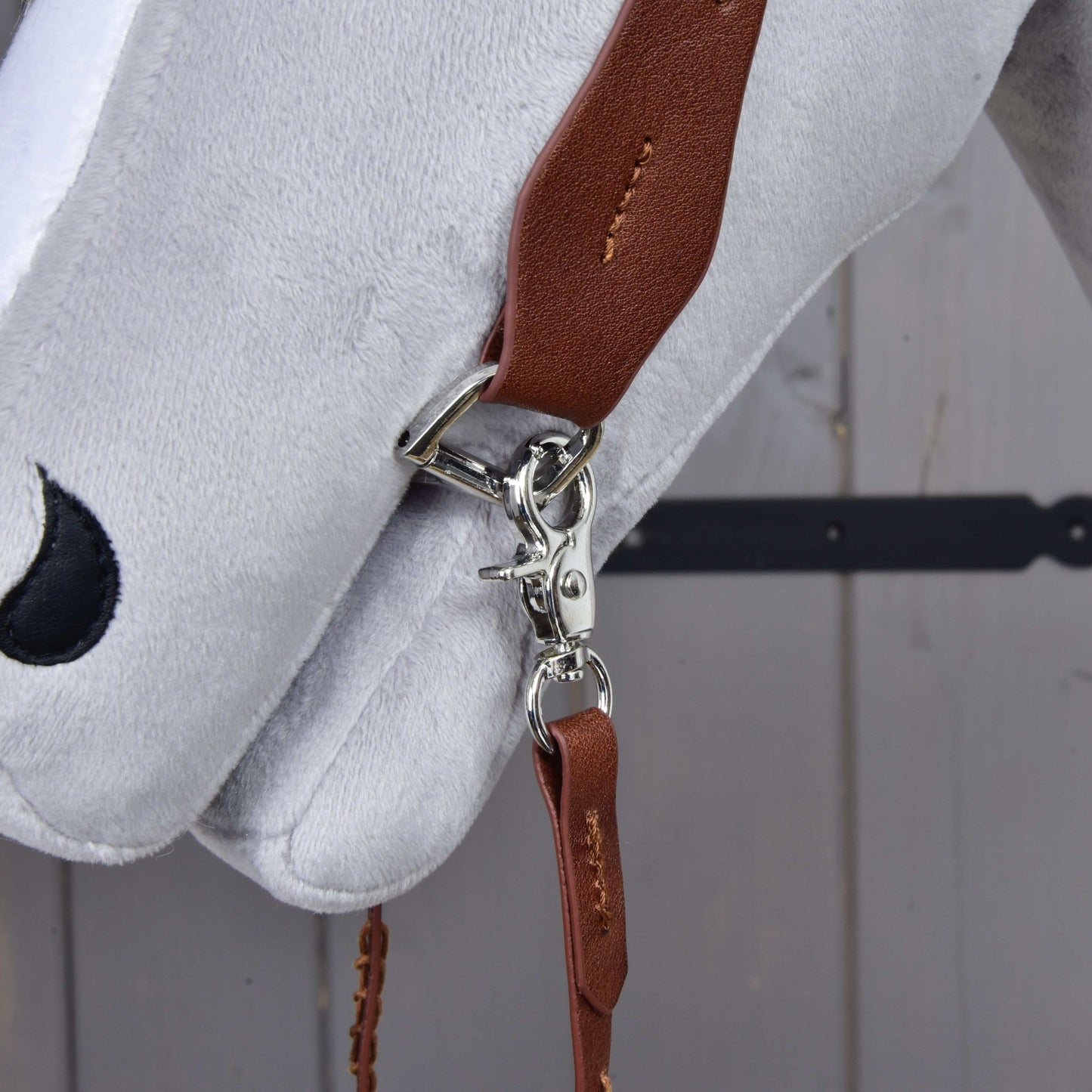A close-up of the Hobby Horse LarDen Complete Western bridle One-Ear, featuring a brown leather strap with a silver clasp and sliding ear loop, attached to a white plush horse's head on a gray wooden background.