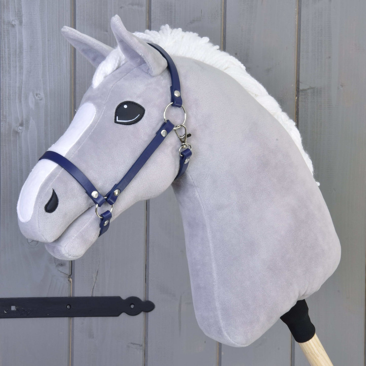 Ein graues Plüsch-Hobby Horse mit weißer Mähne und dem Halfter - dunkelblaues Kunstleder von Hobby Horse LarDen steht auf einem Holzstab vor einer grauen Holzwand.