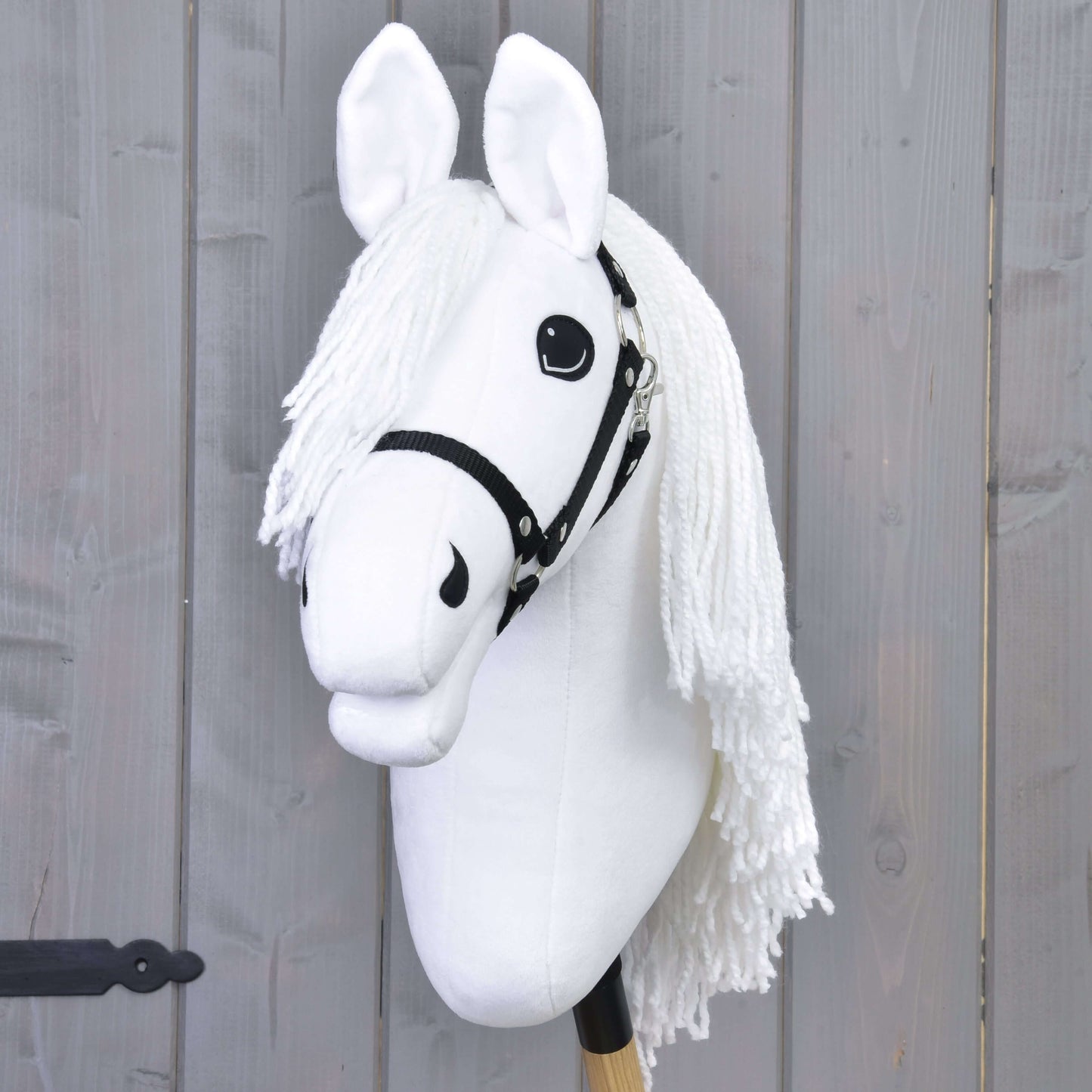 Hobby Horse Lady avec licol noir de Hobby Horse LarDen, avec crinière blanche et yeux expressifs sur bâton en bois devant un fond en planches grises, idéal comme jeu de dressage imaginatif inspiré des chevaux lipizzans.