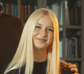 Eine junge Frau mit langen, glatten blonden Haaren lächelt in die Kamera. Sie trägt ein schwarzes Oberteil, mit einem unscharfen Bücherregal im Hintergrund. Eine Hand ruht sanft auf ihrer Schulter und vermittelt einen warmen Moment nach einem schönen Tag mit Hobby Horsing.