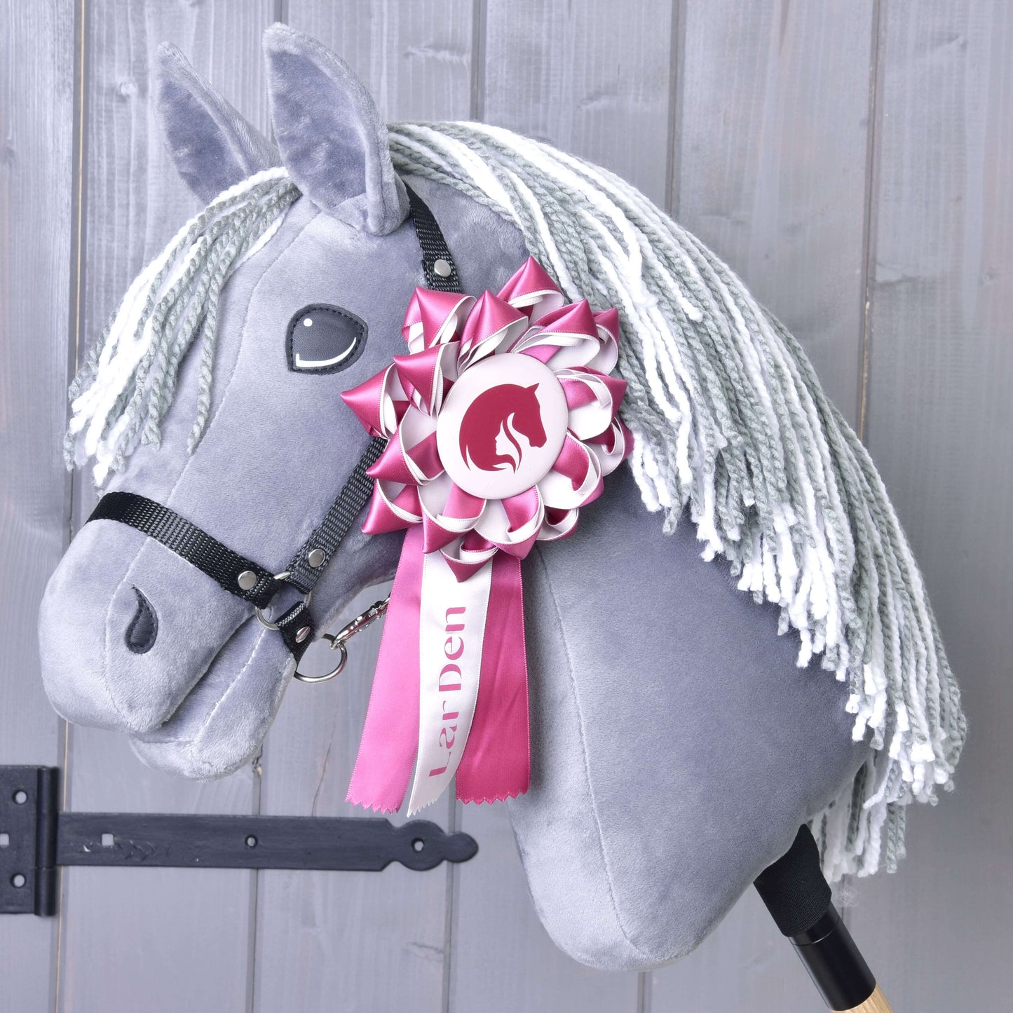 Die Hobby Horse Schleife LarDen Pink von Hobby Horse LarDen ist an einem grauen Hobby Horse mit weißer Garnmähne, schwarzem Zaumzeug und einer rosa-weißen Dressurschleife am Kopf vor hellem Holzhintergrund zu sehen.