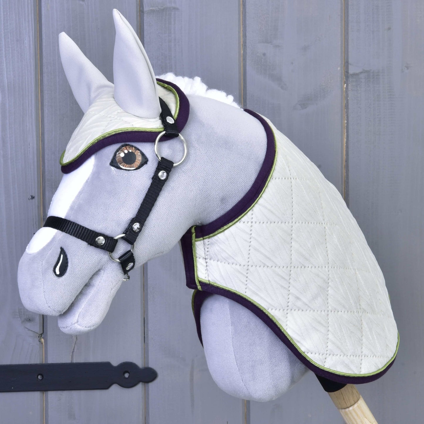 Das Set Leonardo von Hobby Horse LarDen zeigt ein graues Plüsch-Hobby Horse mit weißer gesteppter Decke, passender Fliegenhaube, schwarzem Zaumzeug und Zügeln auf einem Holzstab vor einer grauen Holzwand.