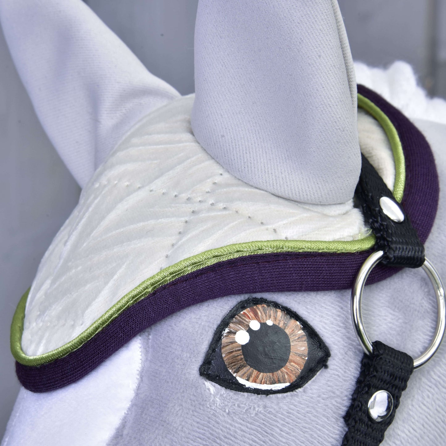 Nahaufnahme von Set Leonardo von Hobby Horse LarDen: ein Stoffpferdekopf mit gestickten Details, einem großen braun-schwarzen Auge, weißer gesteppter Fliegenhaube mit grünem und lila Besatz sowie schwarzem Riemen und silbernem Ring – eine charmante Dekoration.