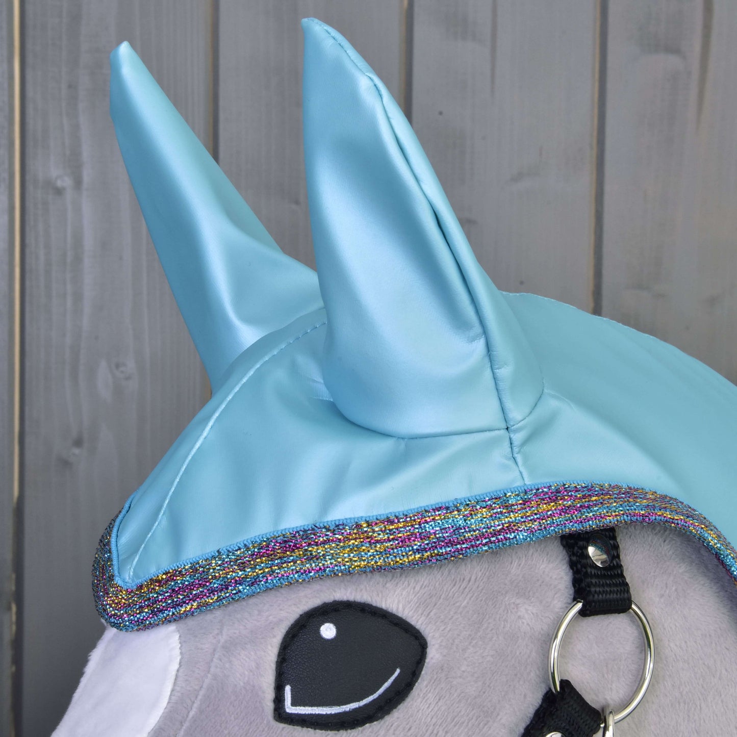 Nahaufnahme der Regendecke Light Blue von Hobby Horse LarDen: ein Plüsch-Einhornkopf mit glänzenden blauen Hörnern, regenbogenfarbiger Einfassung, schwarzem Zaumzeug und gesticktem Auge im Cartoon-Stil – ideal für Spiel bei jedem Wetter vor Holzvertäfelung.