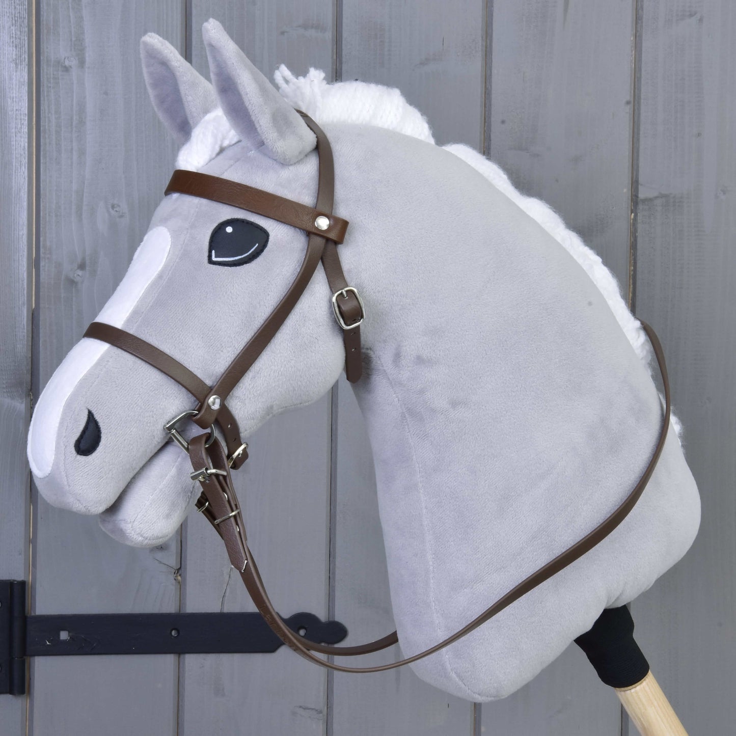 The Hobby Horse LarDen features a gray stick horse toy with a white mane, a friendly expression, and a Complete bridle with noseband Brown, set against a gray wooden background.