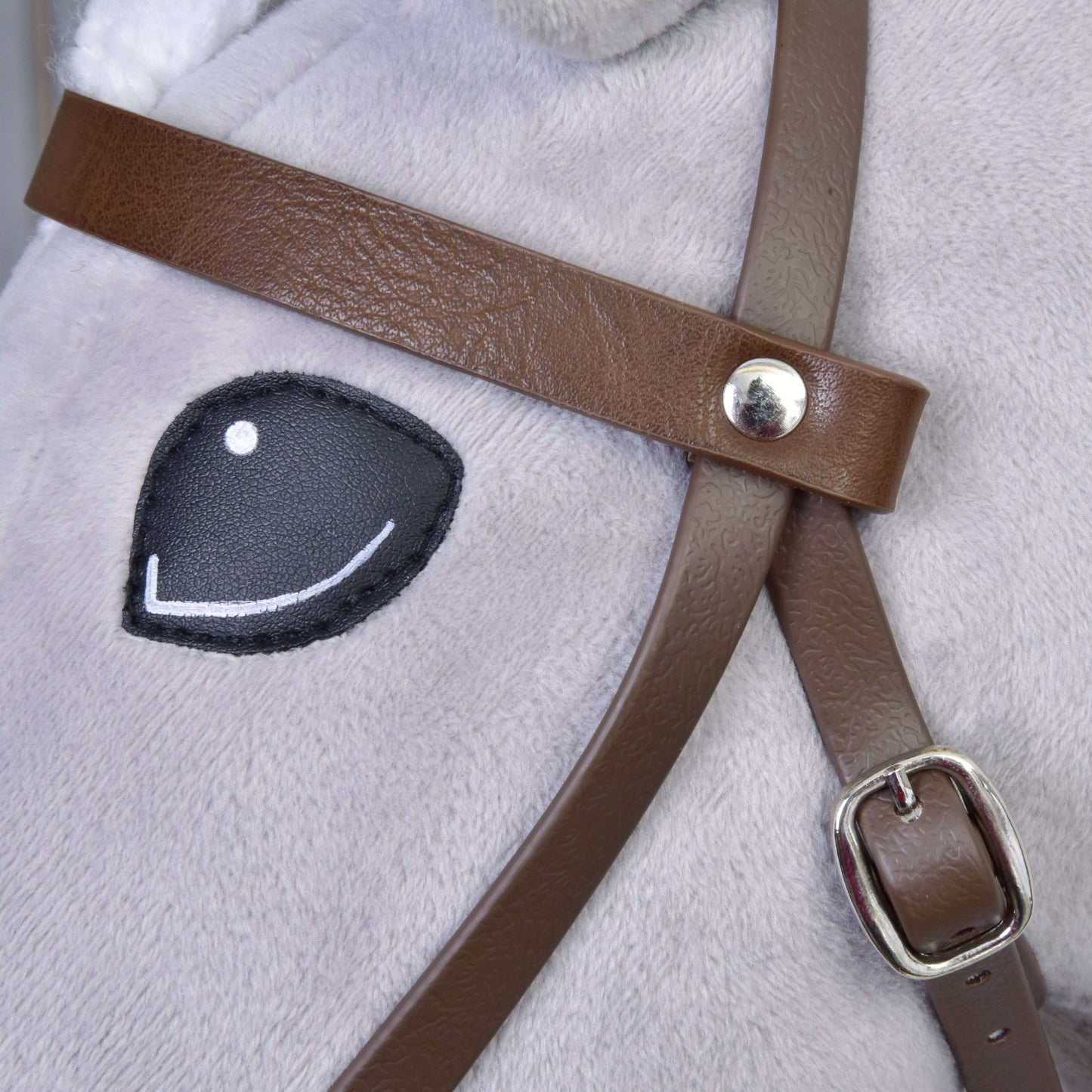 Close-up of a Hobby Horse LarDen plush horse with soft light gray fabric, stitched black eye with white highlight, and a realistic Complete bridle with noseband Brown featuring a metal buckle.