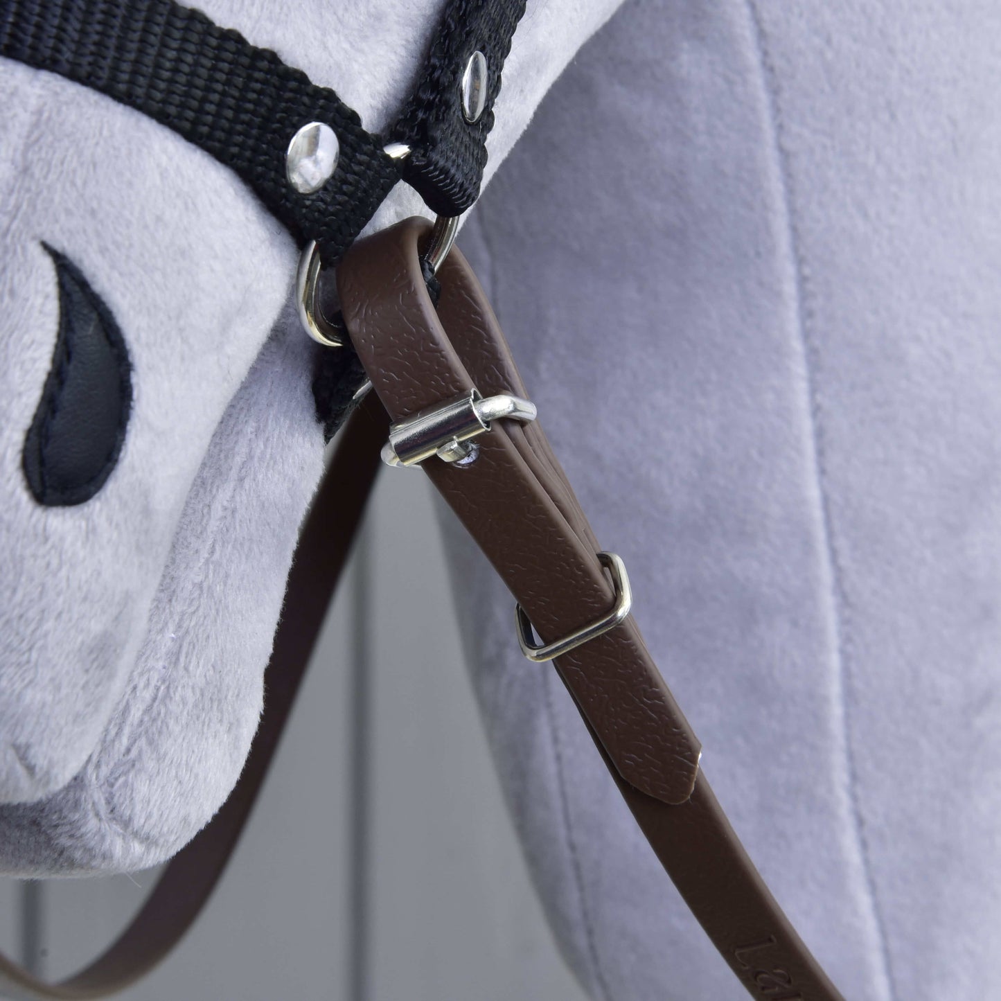 Gros plan sur les rênes marron clair de Hobby Horse LarDen sur un cheval en peluche gris, montrant une sangle en cuir avec boucle, des rênes et une bride en tissu noir, ainsi qu'un œil noir cousu.