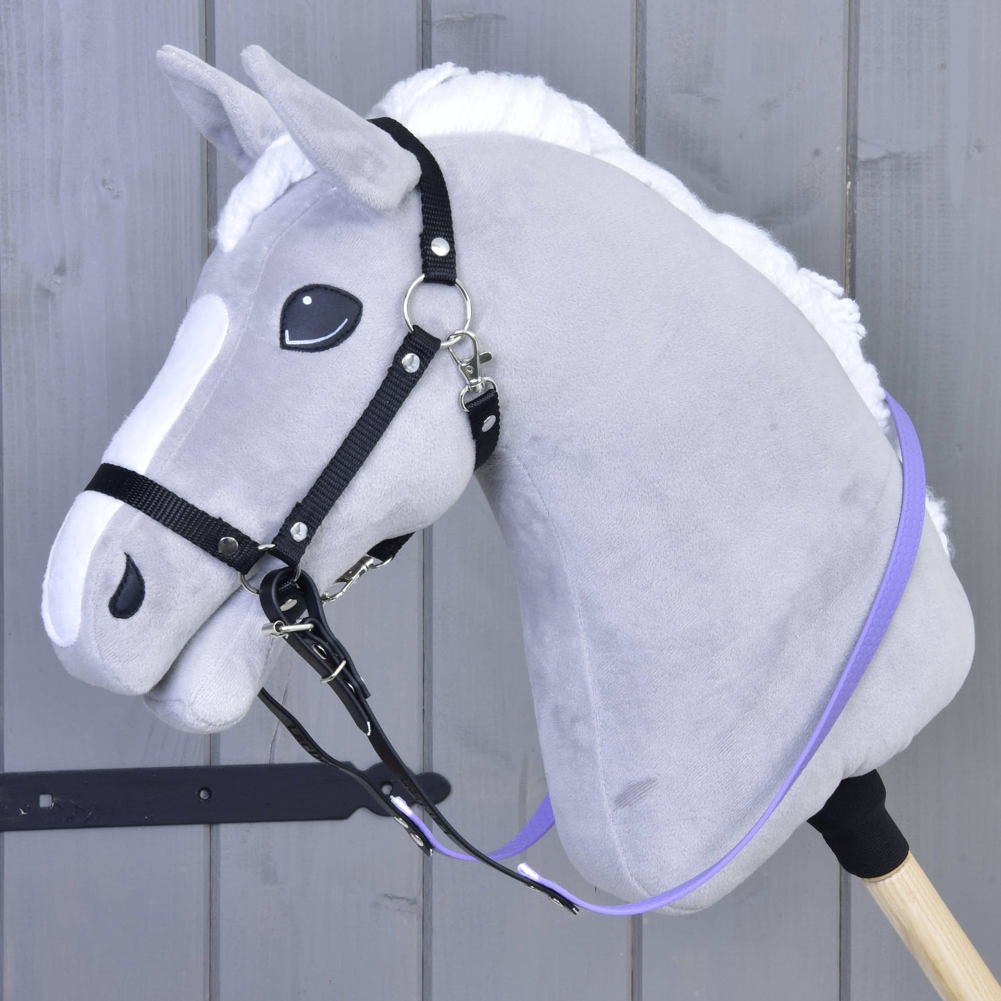 Graues Plüsch-Hobby Horse von Hobby Horse LarDen mit weißer Mähne, gebissloser Zäumung und Gummierte Zügel - Light purple auf einem Holzstab vor Holzpaneel-Hintergrund.