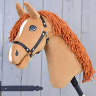Hobby Horse Little Angel mit Schwarzem Halfter von Hobby Horse LarDen mit braunem Plüschkopf, orangefarbener Garnmähne, weißen Abzeichen und schwarzem Halfter, auf einem Stock vor hellgrauem Holzbackground.