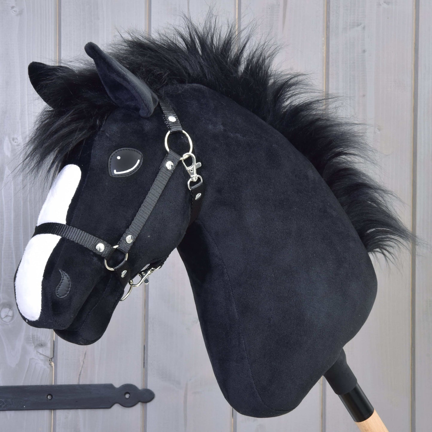 Hobby Horse Loki mit Schwarzem Halfter von Hobby Horse LarDen mit schwarzem Plüschkörper, weißer Blesse und schwarzem Halfter, auf einem Holzstab montiert und vor einer hellen Holzpaneele gezeigt – ideal als Spiel mit Dressurthema.