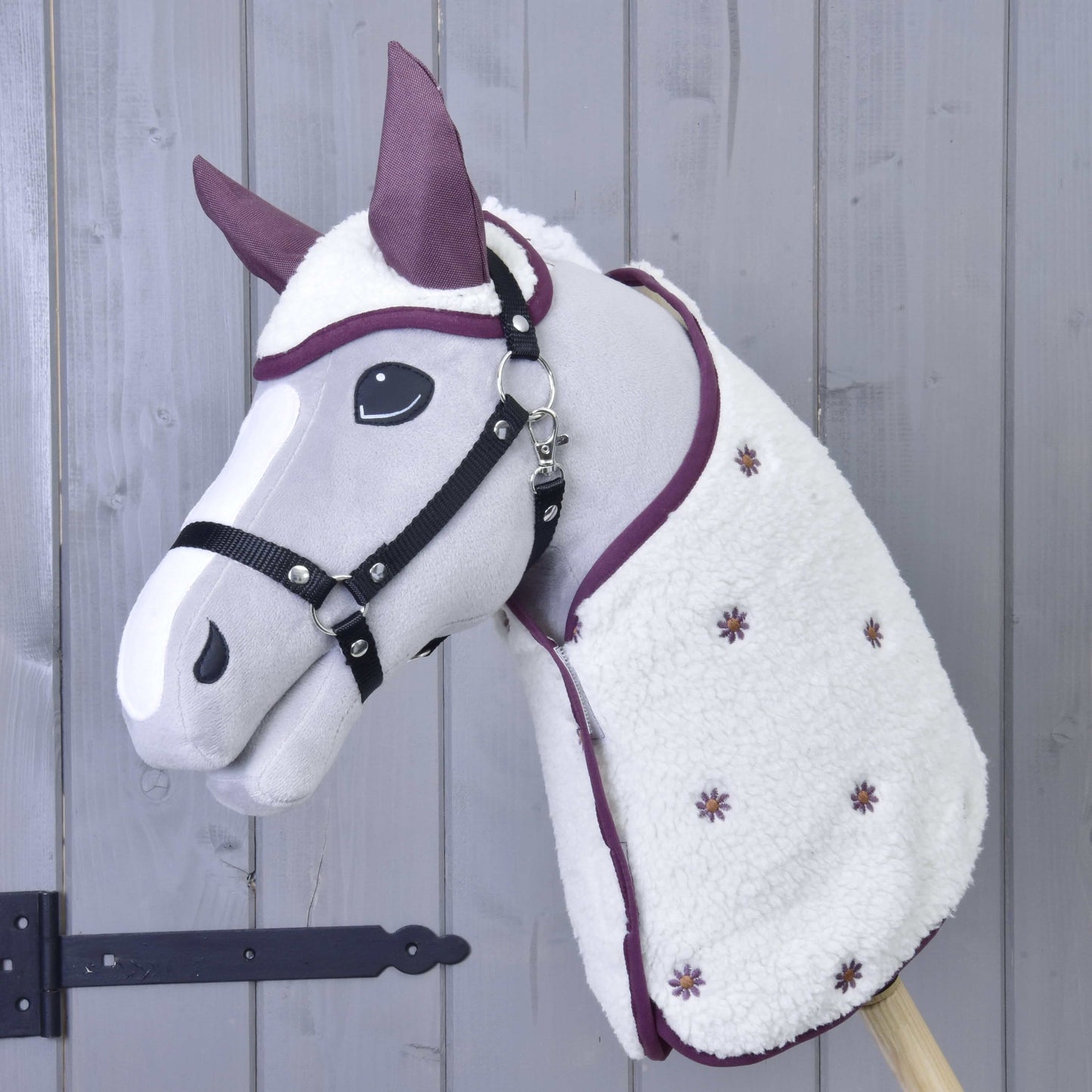 Hobby Horse LarDen Set Loret mit plüschigem Hobby Horse mit grauem Gesicht, schwarzem Zaumzeug, lila Akzenten, Fliegenhaube und weißer Decke mit kleinen lila Blumen, vor einer Wand aus Holzpaneelen.