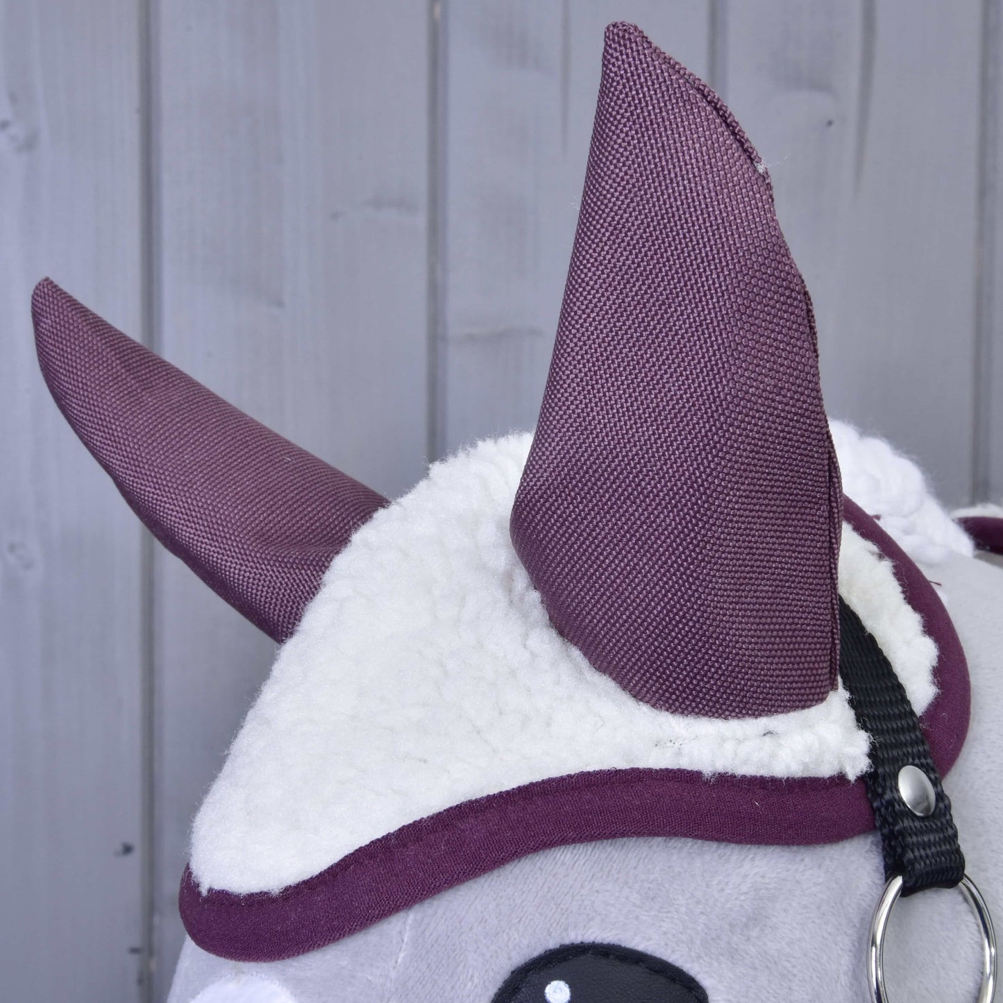 Nahaufnahme des Set Loret von Hobby Horse LarDen, mit weißem flauschigem Stoff, großen lila Ohren im Haubenstil und einem schwarzen Riemen vor einem hellgrauen Holzhintergrund.