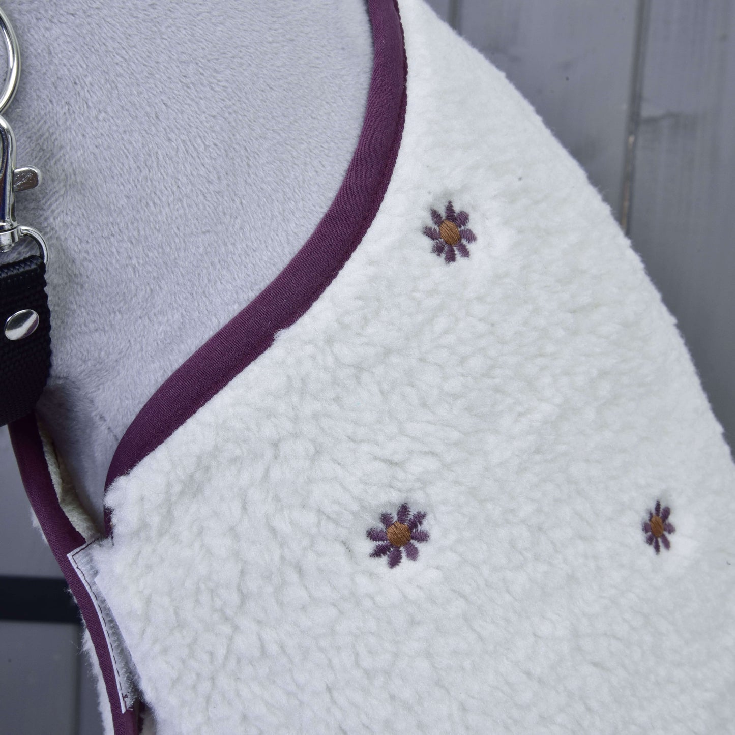 Nahaufnahme von Set Loret, einem weißen Hundemantel aus Fleece von Hobby Horse LarDen, mit lila-gelben gestickten Blumen und lila Besatz. Gezeigt an einem Hund mit schwarzer Leine, ähnelt er einer Decke für Hobby Horse und liegt auf einer grauen Holzoberfläche.