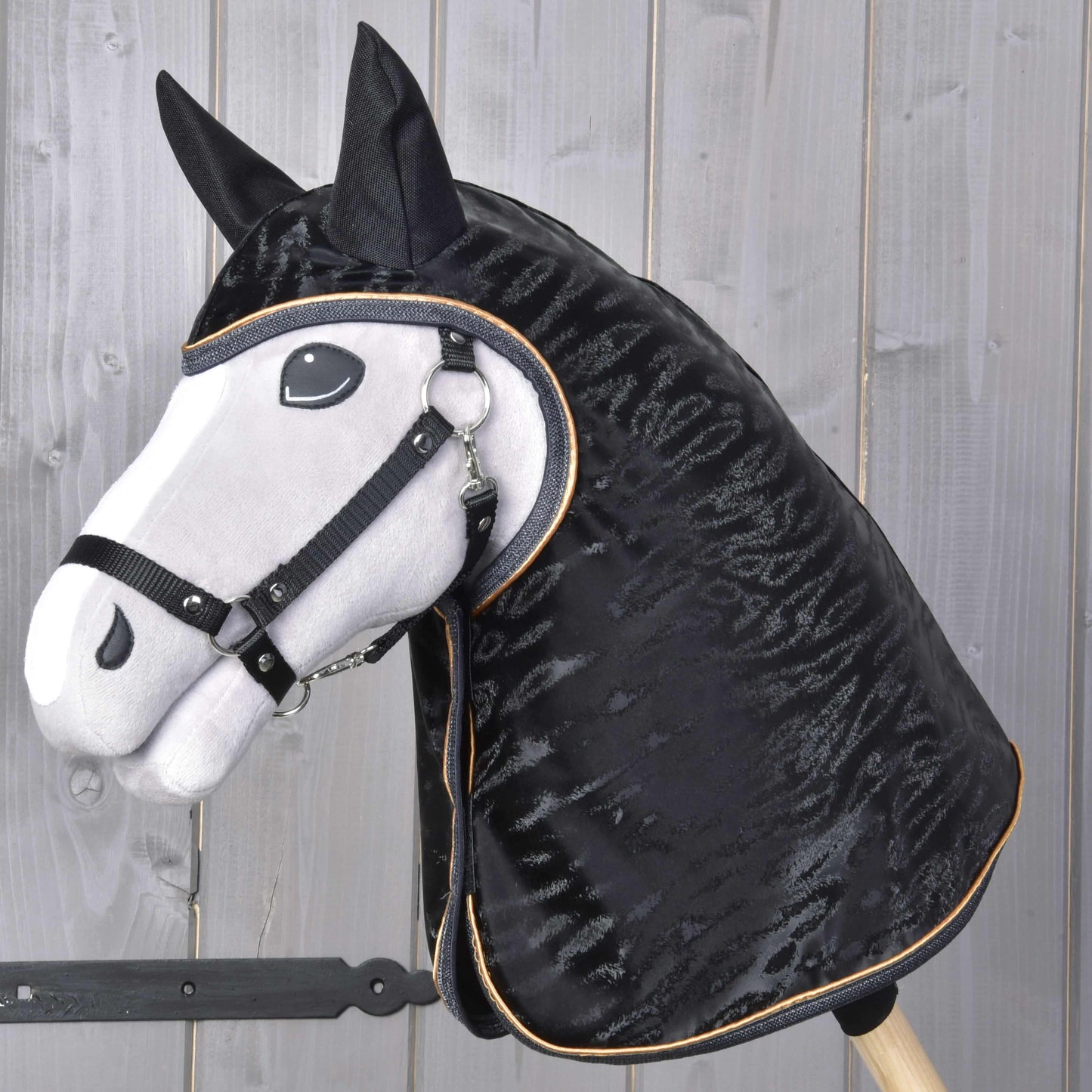 Ein realistisches Hobby Horse mit weißem Gesicht, schwarzer Mähne und schwarzem Zaumzeug steht vor einem Hintergrund aus hellen Holzpaneelen und trägt die Hobby Horse LarDen Regendecke Luxury.
