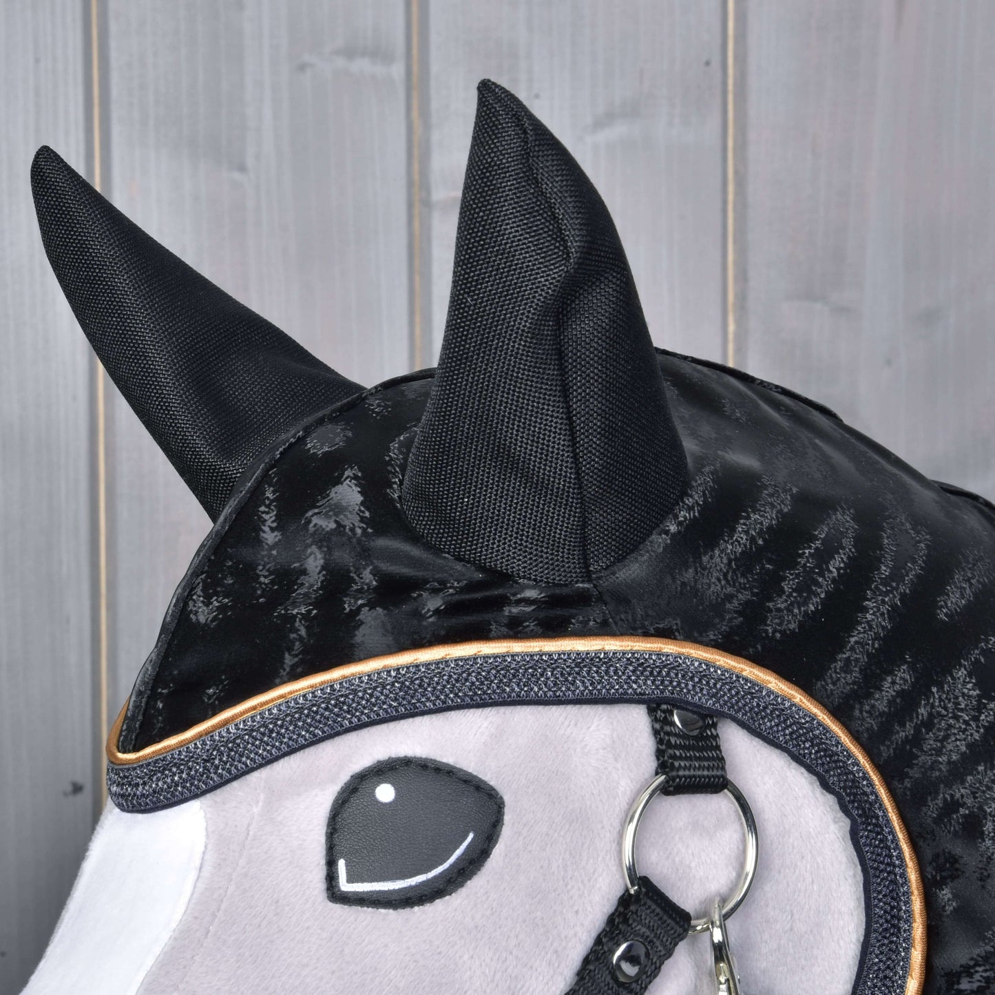 Nahaufnahme der Hobby Horse LarDen Regendecke Luxury: ein schwarz-grauer Stoffkopf eines Hobby Horse mit großen schwarzen Ohren, gesticktem lächelndem Auge, glänzender schwarzer Mähne und einem Metallring am Maul vor Holzpaneelen.