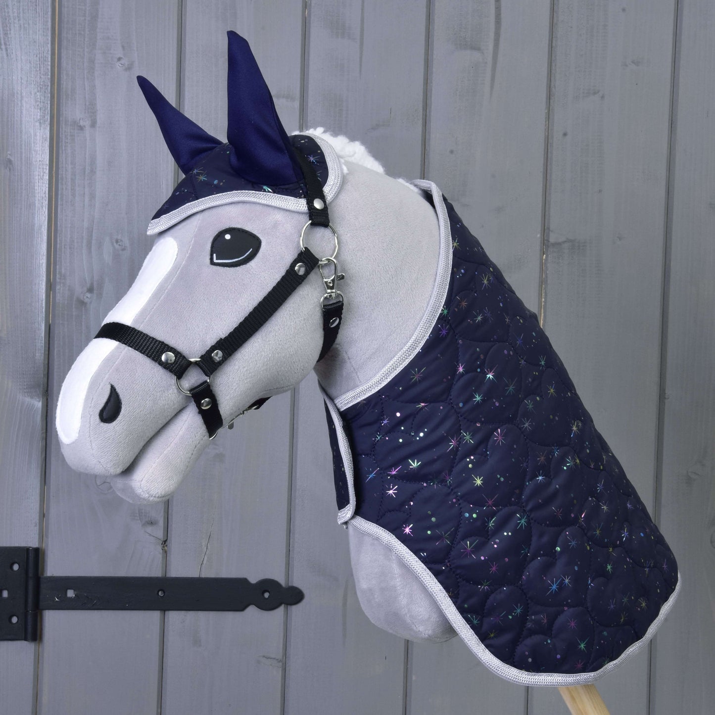 Hobby Horse LarDen Set Magic mit plüschigem Steckenpferd, dunkelblauer gesteppter Decke und Fliegenhaube mit Sternenmuster als Insektenschutz, vor einer Holzwand.