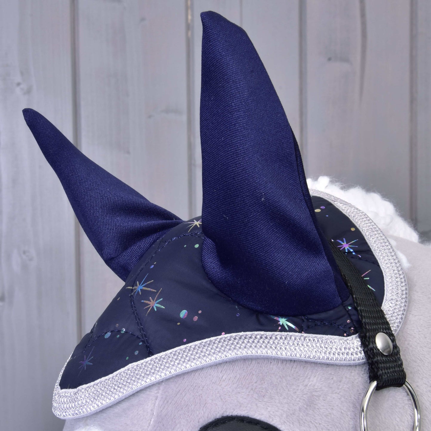 Nahaufnahme von Hobby Horse LarDen Set Magic: ein plüschiger Pferdekopf mit blauer Fliegenhaube mit funkelndem Sternenmuster und weißem Besatz als Insektenschutz, vor hellem Holz-Hintergrund.