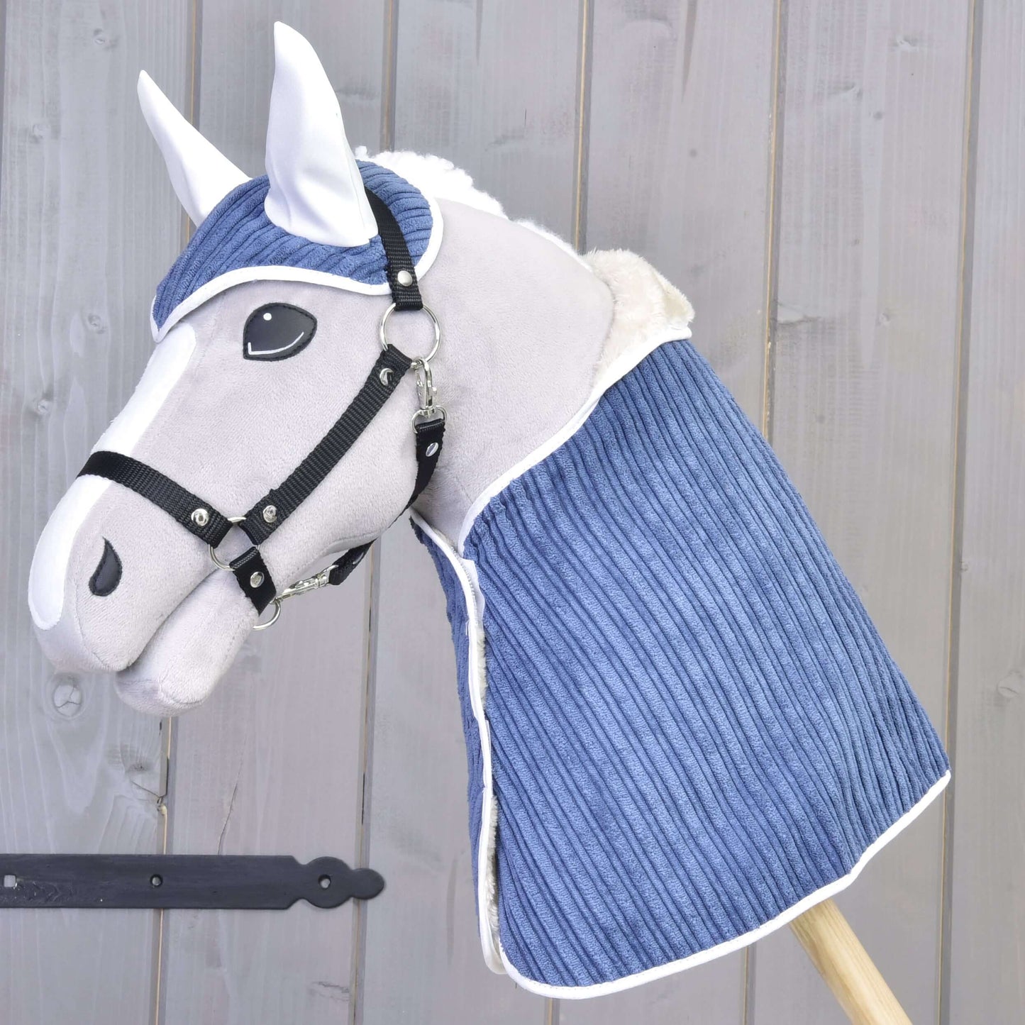 Das Set Manchester Plum von Hobby Horse LarDen zeigt ein Plüsch-Hobby Horse mit weißem Gesicht, schwarzem Zaumzeug und blauer gesteppten Decke sowie passender Fliegenhaube, vor einer hellen Holzwand.