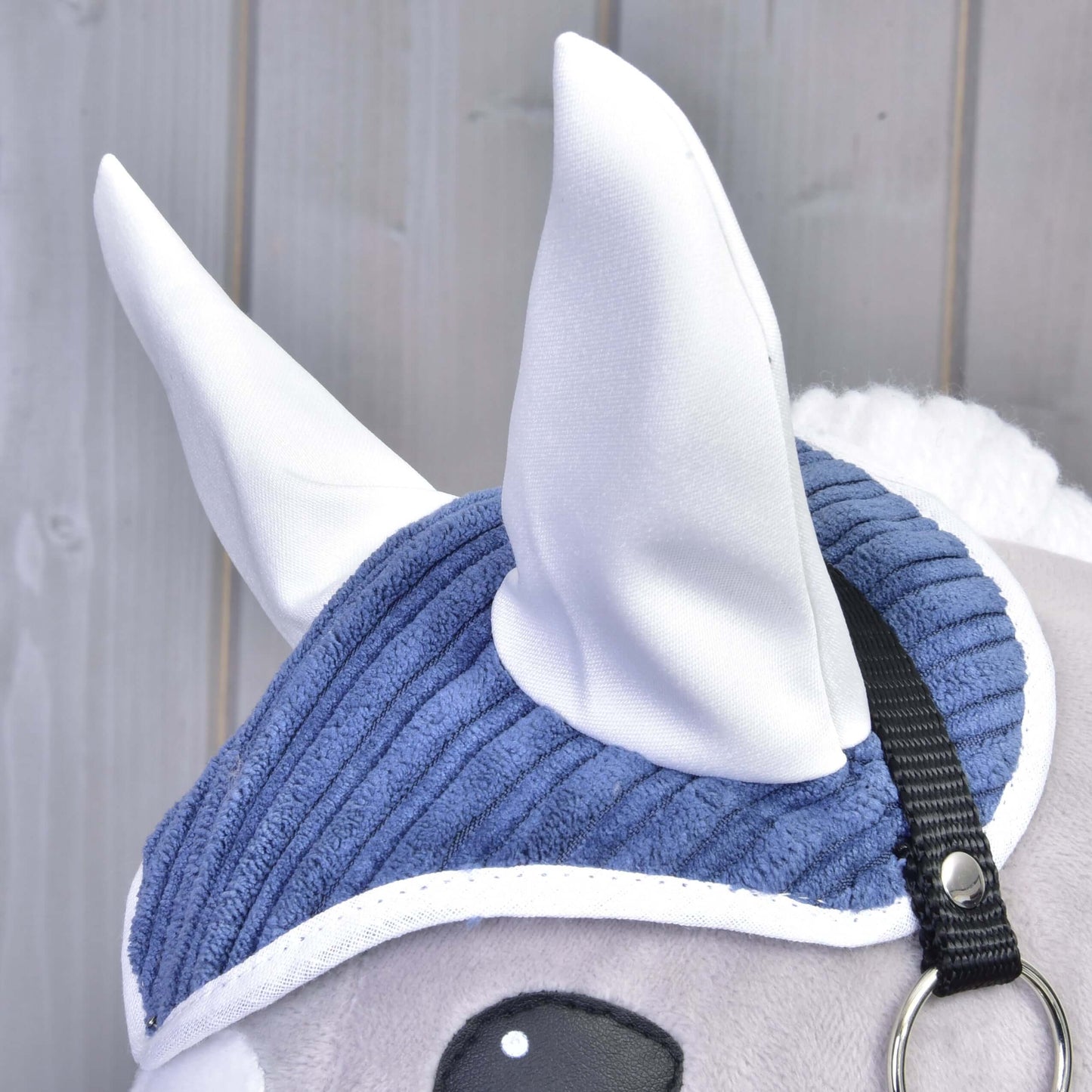 Nahaufnahme des Set Manchester Plum von Hobby Horse LarDen mit weißen Stoffohren, blauer Cord-Fliegenhaube, schwarzem Riemen und weißer Mähne vor hellem Holzbackground.