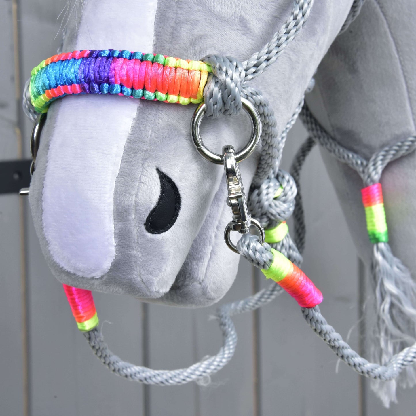 Gros plan sur une tête de Hobby Horse grise en peluche de Hobby Horse LarDen, avec le licol arc-en-ciel et les rênes en corde grise de l’ensemble de cordes Manhattan, narine noire cousue, sur fond de panneaux en bois clair.