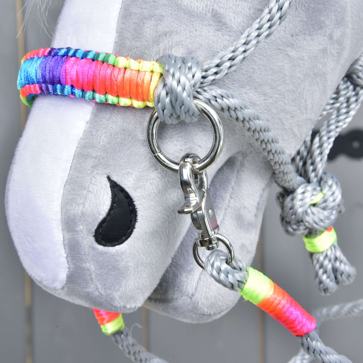 Gros plan sur l’ensemble de cordes Manhattan de Hobby Horse LarDen, avec tête de Hobby Horse grise rembourrée, licol tressé coloré, anneau métallique, clip et rênes assorties sur fond en bois clair.