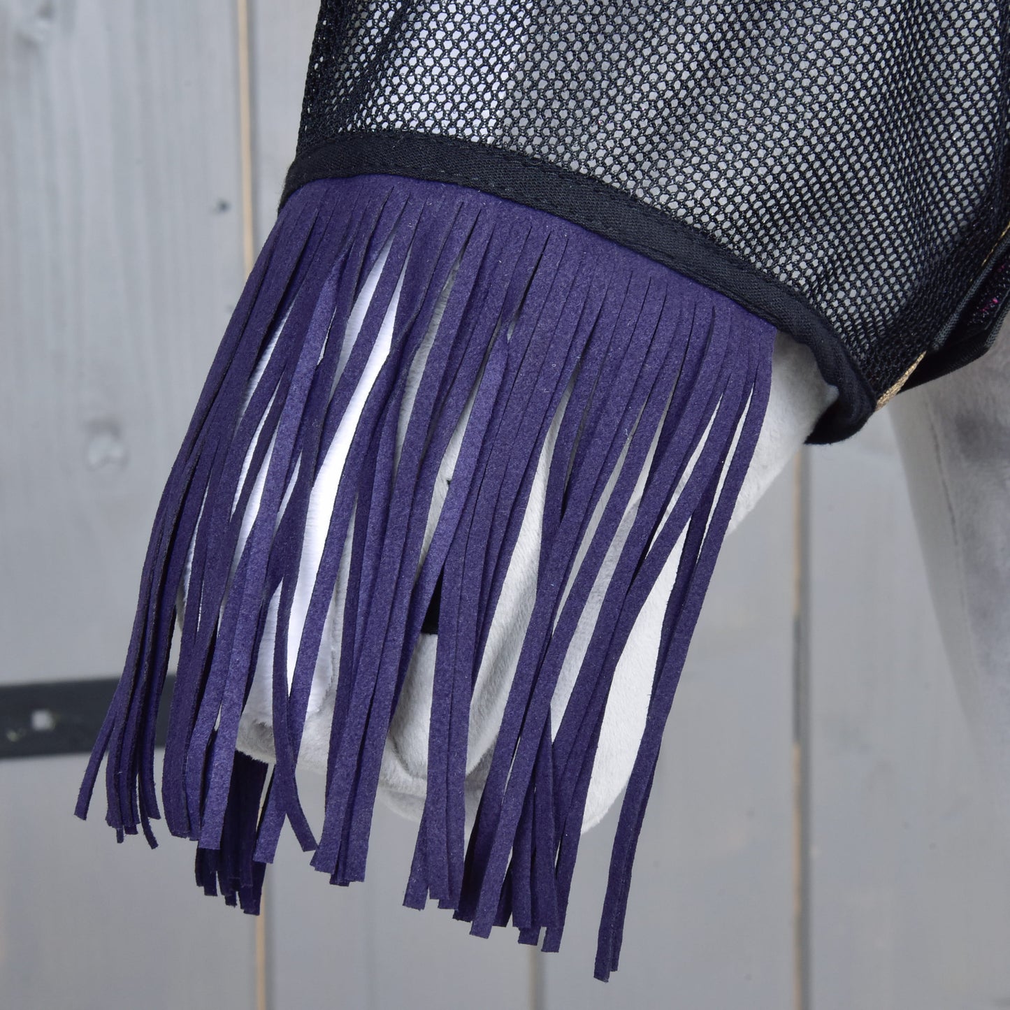 Gros plan sur un cheval portant le Masque anti-mouches De Luxe Mona de Hobby Horse LarDen, avec maille noire et longues franges violet foncé, sur fond en bois clair.