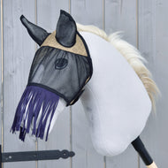 Fliegenmaske de Luxe Mona von Hobby Horse LarDen auf einem weißen Plüsch-Hobby Horse mit schwarzem Stabgriff und hellblonder Mähne, mit stilvoller Fransen-Fliegenmaske vor grauem Holzhintergrund.
