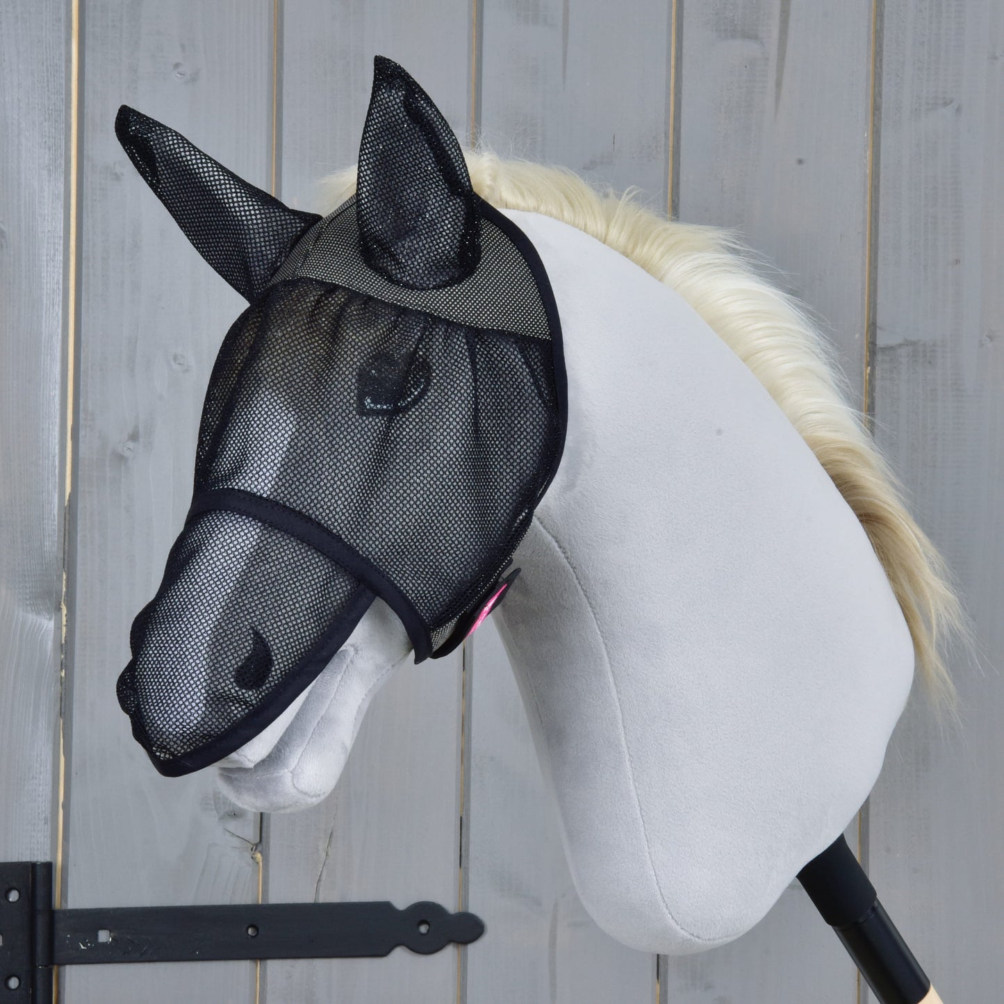 Ein weißes Hobby Horse mit heller Mähne trägt die Hobby Horse LarDen Fliegenmaske de Luxe Full, sein Gesicht durch schwarzes Netz geschützt, vor einem Holz-Hintergrund.