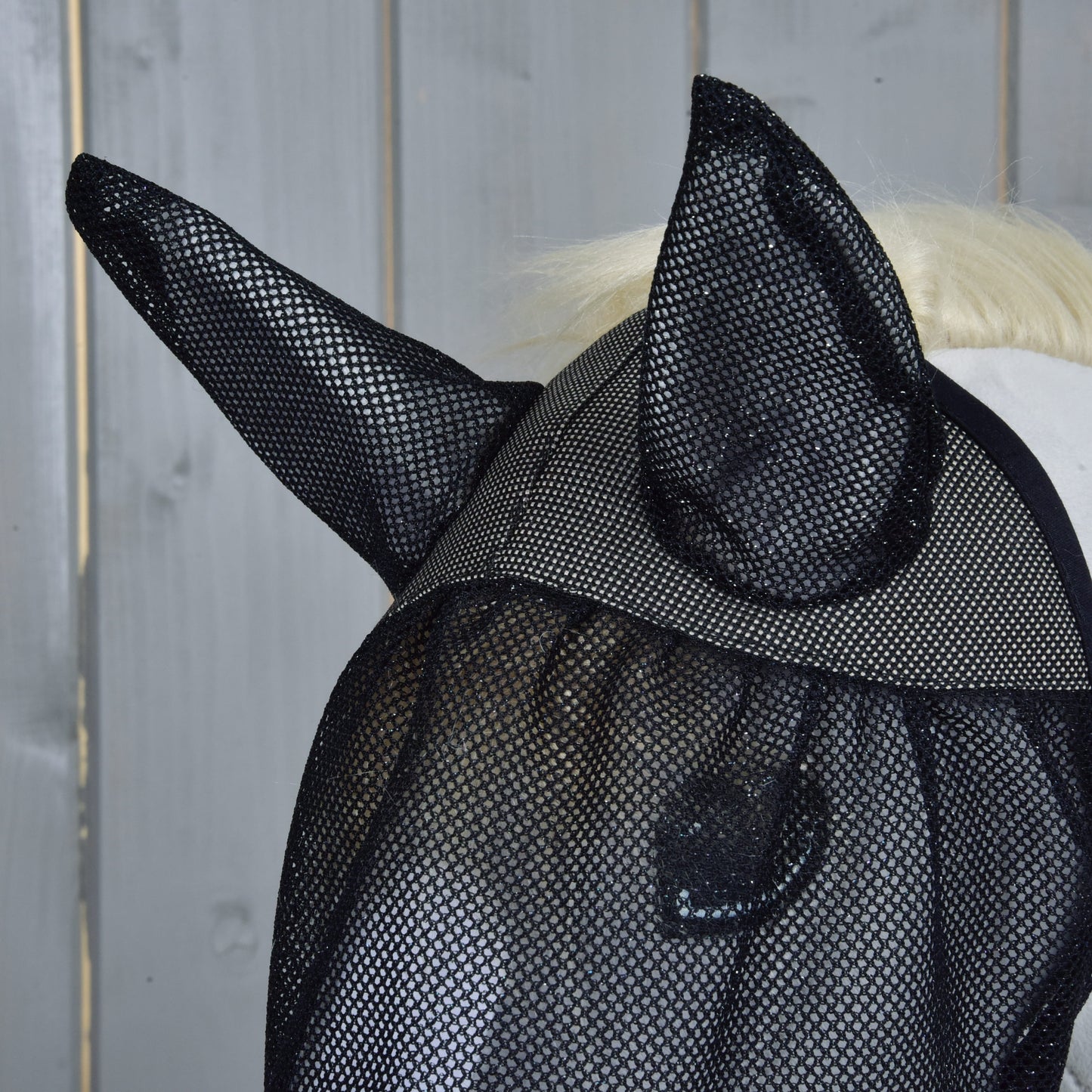 Nahaufnahme eines Pferdes mit der Hobby Horse LarDen Fliegenmaske de Luxe Full mit Ohrenschutz, vor einem grauen Holz-Hintergrund.