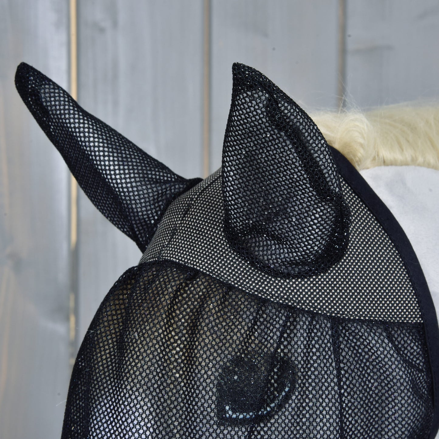 Nahaufnahme eines Pferdekopfes mit der Fliegenmaske de Luxe Full von Hobby Horse LarDen zum Schutz, mit einer Holzwand im Hintergrund.