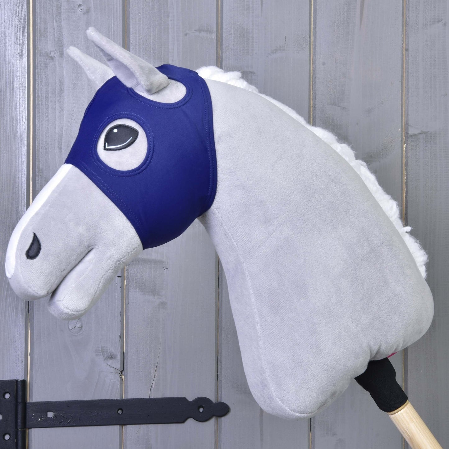 Masque de cheval sans oreilles LarDen de Hobby Horse LarDen sur un Hobby Horse gris avec crinière blanche et manche en bois, contre un mur en bois avec charnières visibles.