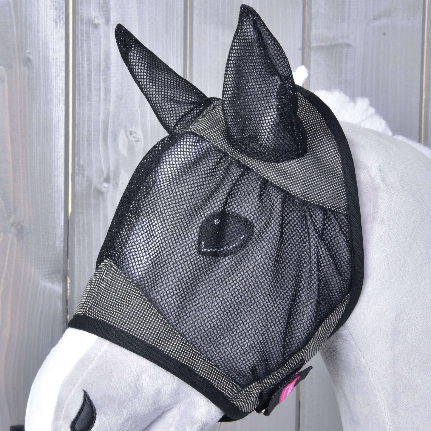 Cheval blanc près d’une clôture en bois portant le Masque anti-mouches De Luxe Gris de Hobby Horse LarDen, un masque lavable avec couvre-oreilles qui protège les yeux, les oreilles et une partie du visage des insectes.