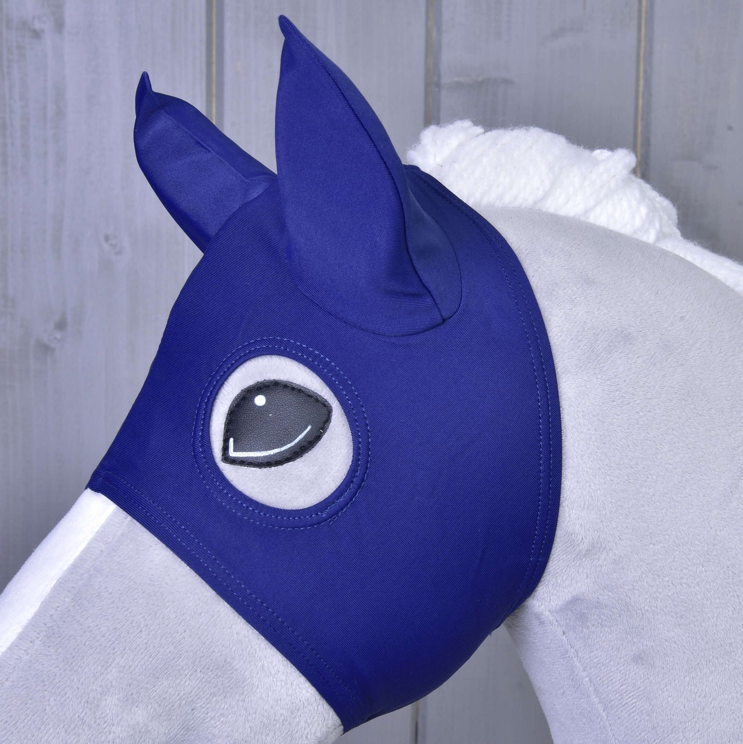 Gros plan d’un cheval gris portant le Masque de cheval LarDen de Hobby Horse LarDen, avec couvre-oreilles et ouvertures pour les yeux, devant un fond en bois.
