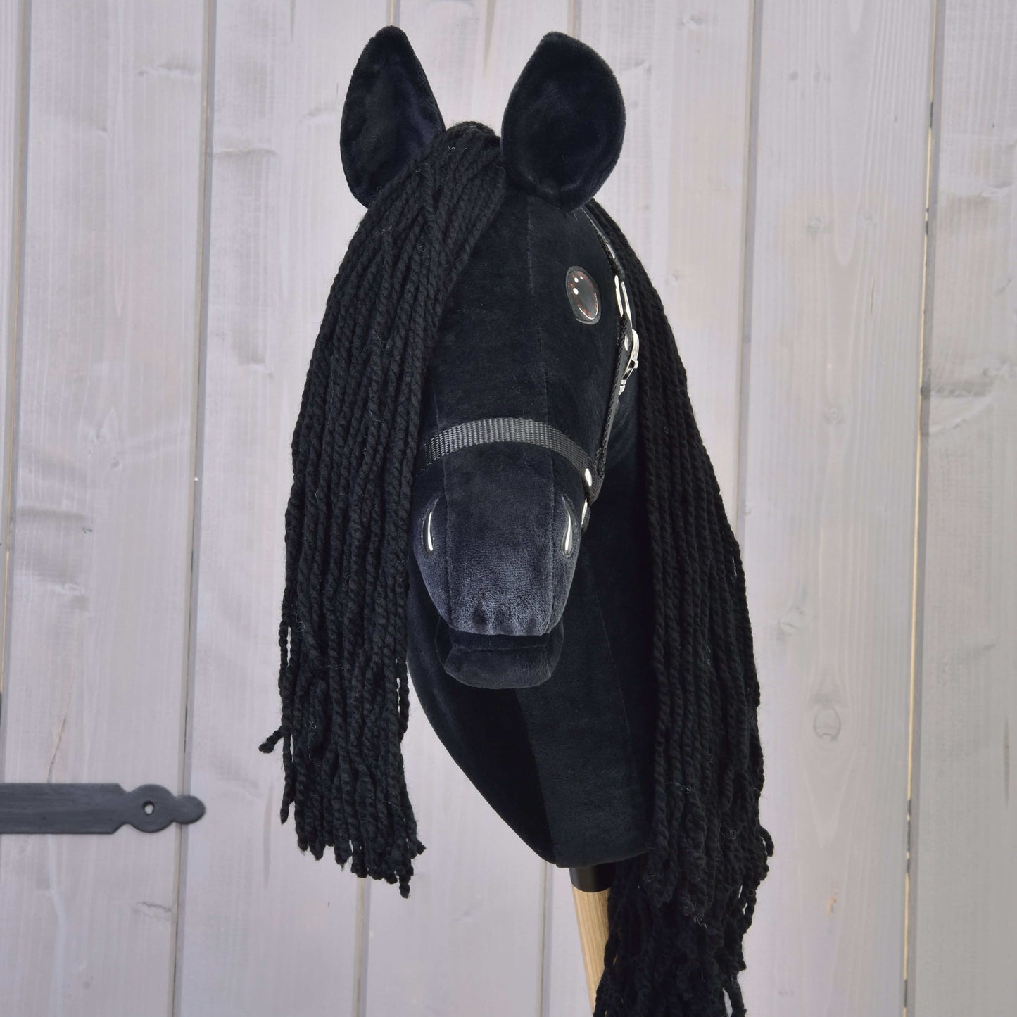 Hobby Horse Maximus avec licol noir de Hobby Horse LarDen, Hobby Horse frison fait main avec tête en peluche, crinière en fil, yeux en bouton, licol noir et bâton en bois, présenté sur fond bois clair.