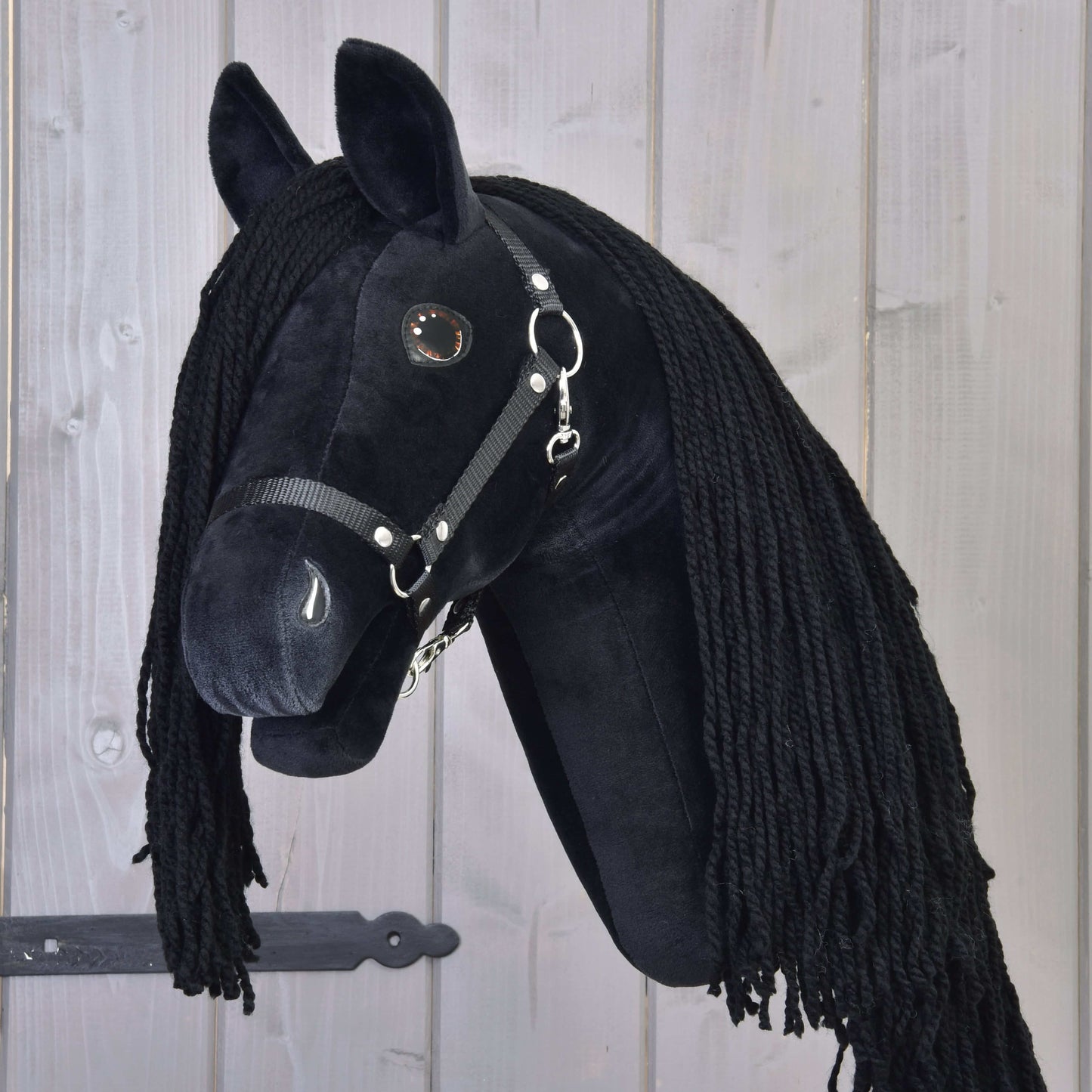 Hobby Horse Maximus avec licol noir de Hobby Horse LarDen, Hobby Horse frison noir en peluche avec longue crinière noire, monté sur un bâton en bois et présenté devant un fond en panneaux de bois clair.