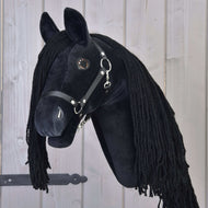 Hobby Horse LarDen Hobby Horse Maximus mit Schwarzem Halfter, ein plüschiges schwarzes Friesisches Hobby Horse mit langer schwarzer Mähne auf einem Holzstab, vor einem Hintergrund aus hellen Holzpaneelen.