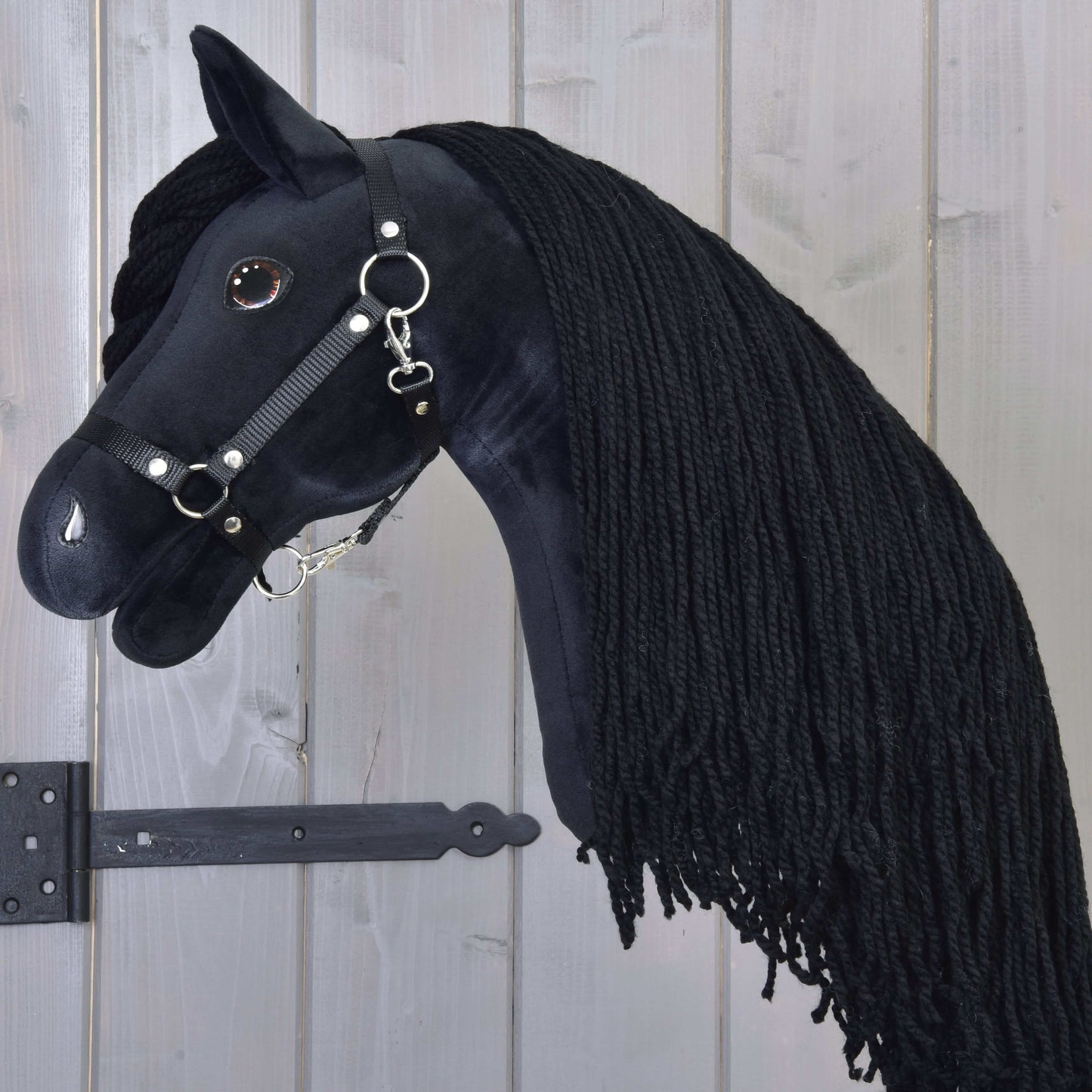 Hobby Horse Maximus avec licol noir de Hobby Horse LarDen, avec design frison noir en peluche, longue crinière tressée, bride avec anneaux en métal et œil brillant en bouton, suspendu sur un mur en bois.