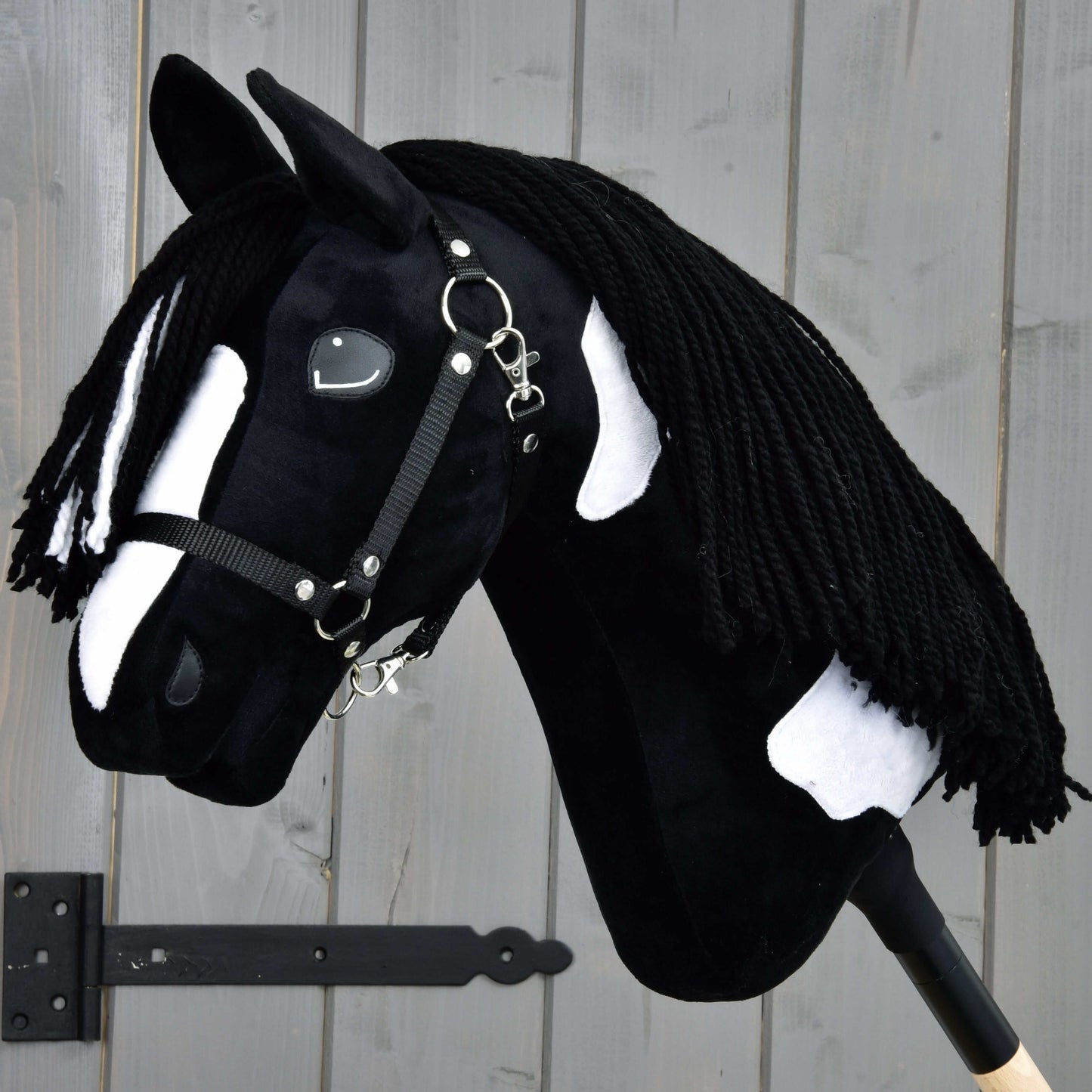 Hobby Horse Merlin avec licol noir de Hobby Horse LarDen, inspiré d’un étalon Barock Pinto, avec taches noires et blanches et crinière tressée en fil, monté devant un mur en bois gris.
