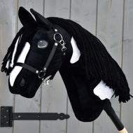 Hobby Horse LarDen „Hobby Horse Merlin mit Schwarzem Halfter“, inspiriert von einem Barock Pinto Hengst, zeigt schwarze und weiße Flecken und eine geflochtene Garnmähne, montiert vor einer grauen Holzwand.