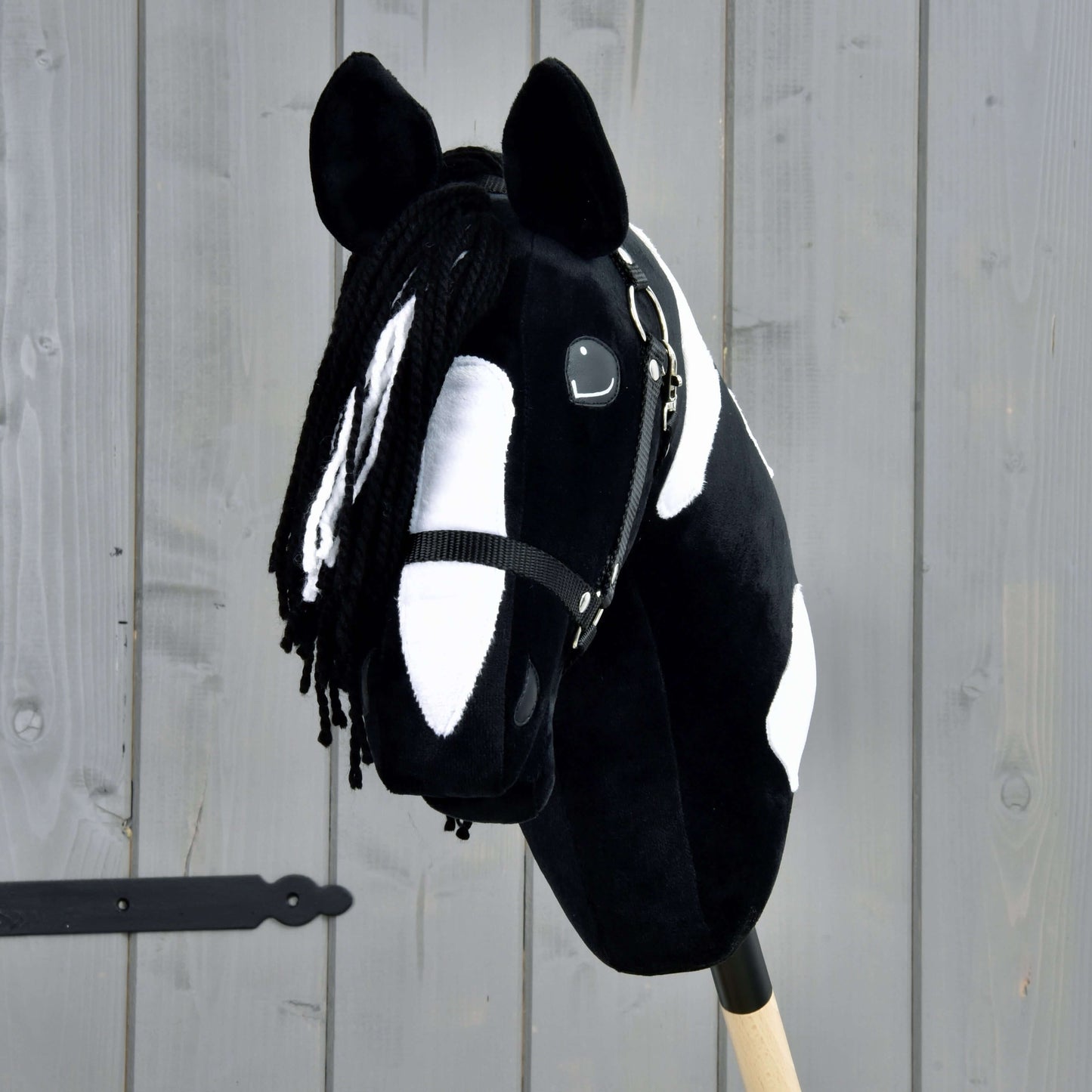 Hobby Horse Merlin avec licol noir de Hobby Horse LarDen, Hobby Horse tacheté noir et blanc inspiré d’un étalon Barock Pinto, avec crinière en fil et bride, monté sur un bâton en bois devant une clôture grise.