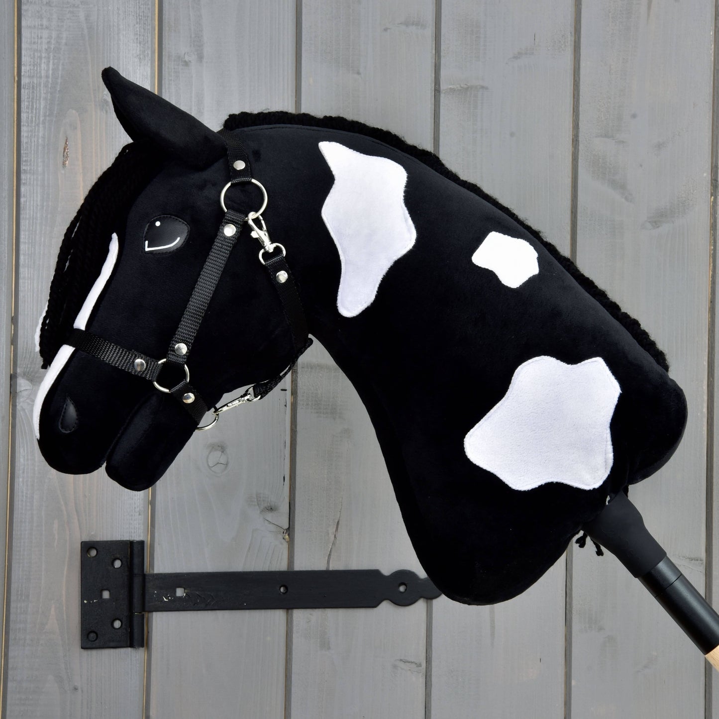 Hobby Horse Merlin avec licol noir de Hobby Horse LarDen, peluche d’étalon Barock Pinto noir et blanc avec visage souriant et crinière noire, présentée montée sur un mur en bois.