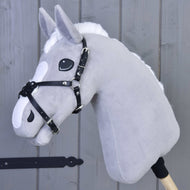 Hobby Horse LarDen Reithalfter Mexico schwarz gepolstert an einem grauen Plüsch-Hobby Horse mit weißer Mähne, schwarzem Zaumzeug und Reithalfter aus Kunstleder auf einem Holzstab vor grauem Hintergrund mit Holzpaneelen.