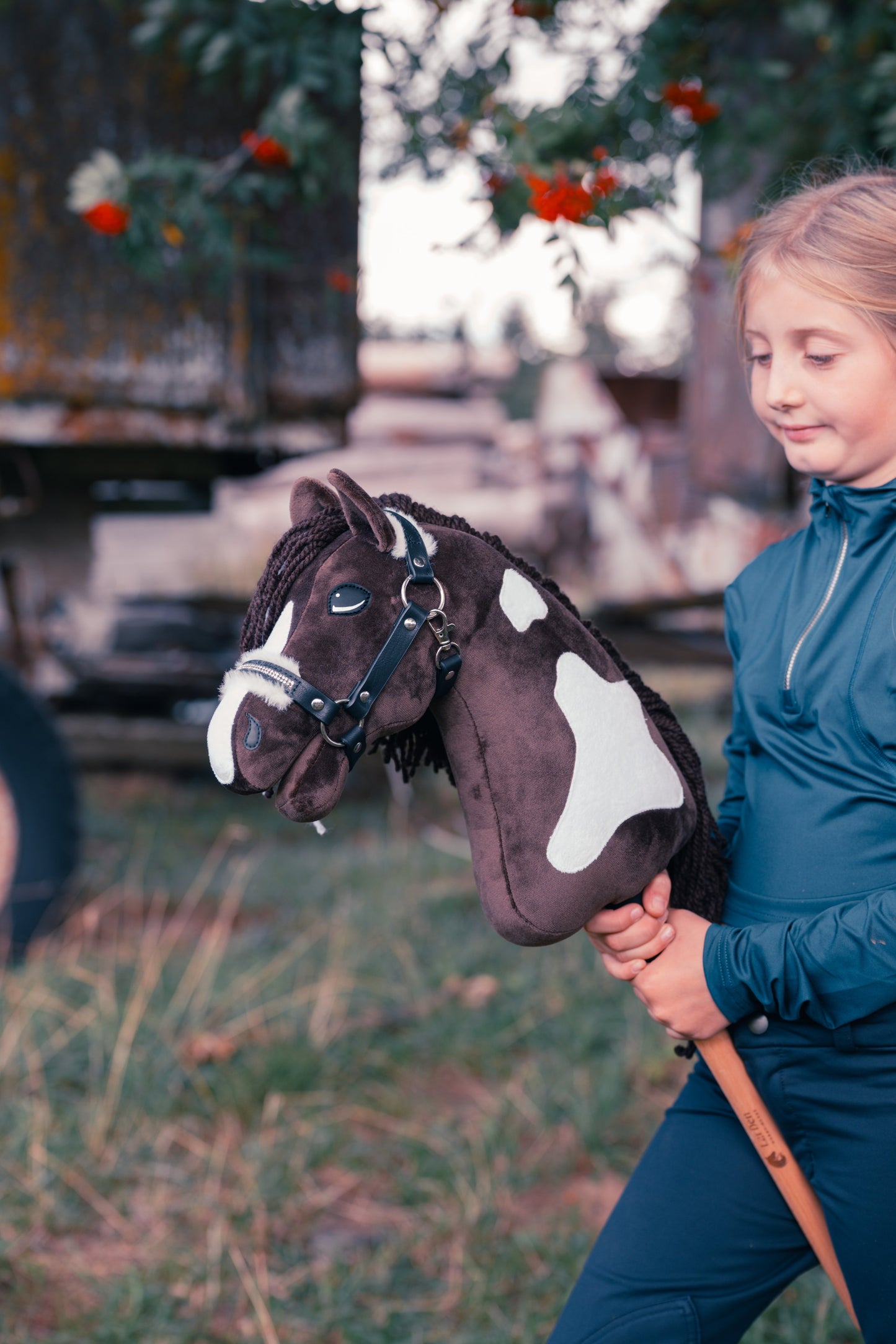 Lifestyle photo: a young rider in a dark blue riding outfit holds the Hobby Horse Monte by Hobby Horse LarDen in a meadow in front of an old wooden wagon and a rowan tree with red berries - brown and white coat inspired by the American Paint Horse, long dark yarn mane, shown here with the fur-lined Espresso halter.