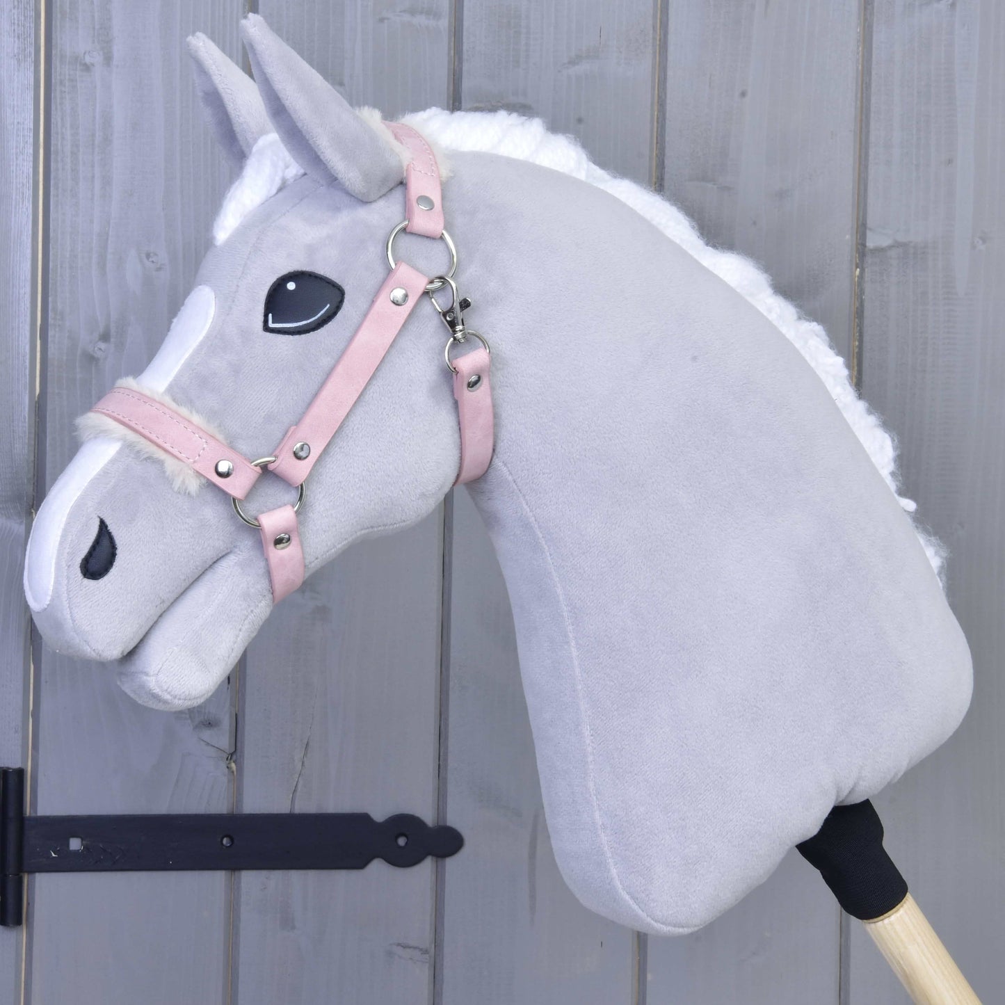 Licol avec fourrure - similicuir Mulberry de Hobby Horse LarDen, Hobby Horse en peluche gris clair avec crinière blanche, bride en similicuir rose et détails brodés, monté sur un bâton en bois devant un mur en bois gris.
