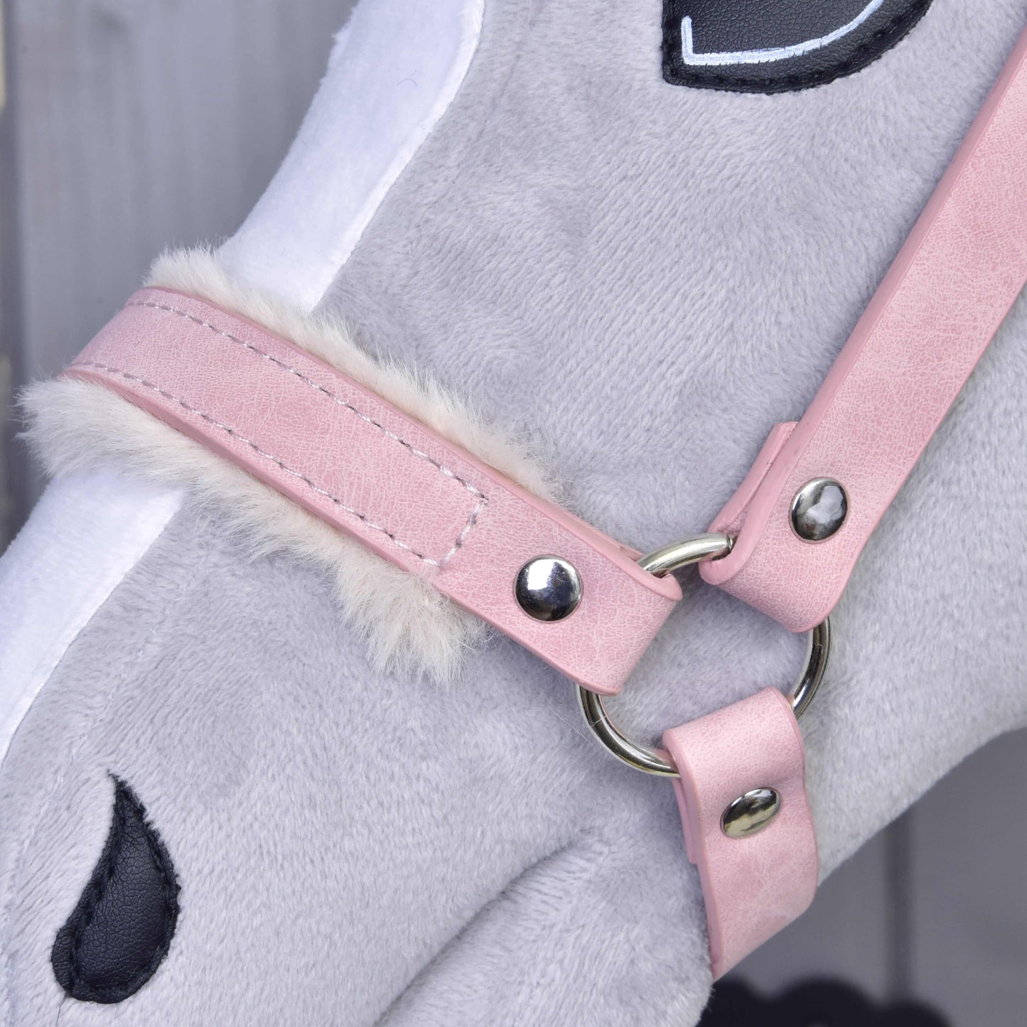 Gros plan sur un cheval jouet en peluche grise portant le licol avec fourrure - similicuir Mulberry de Hobby Horse LarDen, avec similicuir rose clair, bordure en fausse fourrure blanche et détails brodés noirs pour l’œil et le naseau.