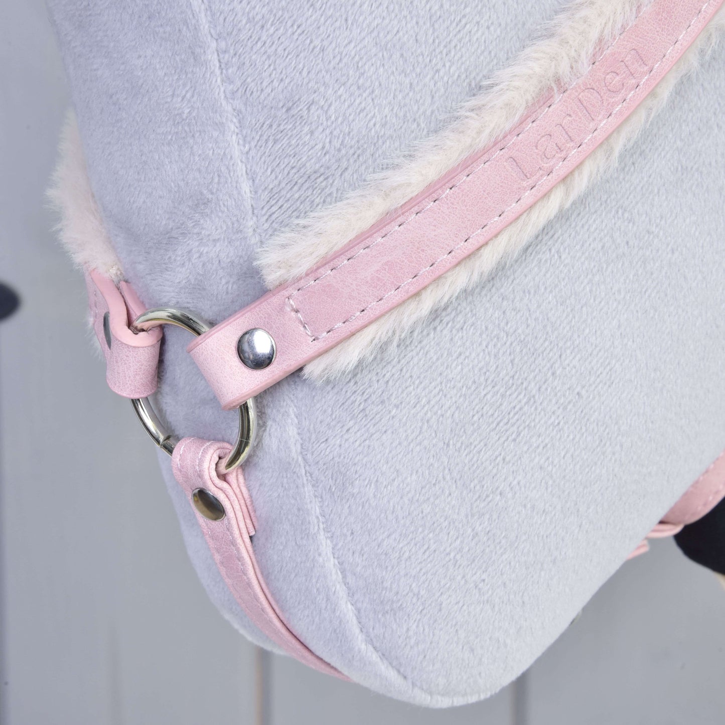 Close-up of the Hobby Horse LarDen Breastgirth with fur Mullbery, featuring light grey plush, a pink leatherette strap with embossed text, white faux fur trim, and metal ring. The background is blurred.