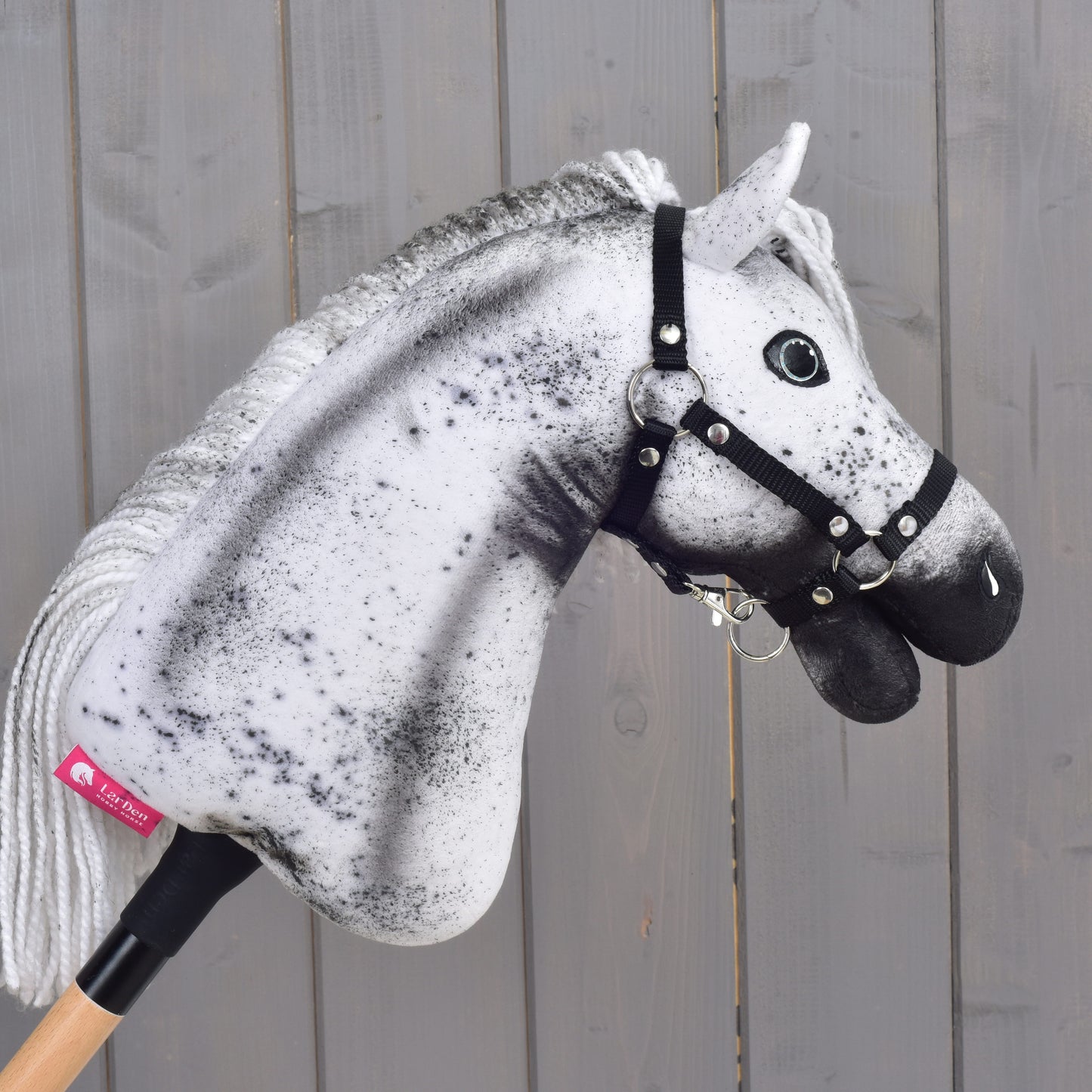 Hobby Horse Naivy avec licol noir - taille S de Hobby Horse LarDen, avec un design moucheté blanc et gris remarquable, fièrement monté sur un bâton en bois devant un mur en bois gris.
