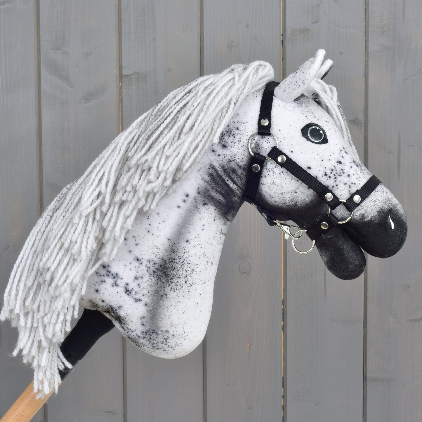 Hobby Horse Naivy avec licol noir - taille S de Hobby Horse LarDen : un Hobby Horse Appaloosa fait main, avec une crinière en fil blanc et gris, des taches noires marquées et un élégant licol noir, placé devant un fond en bois rustique. Idéal comme compagnon pour des aventures imaginaires d’étalon.
